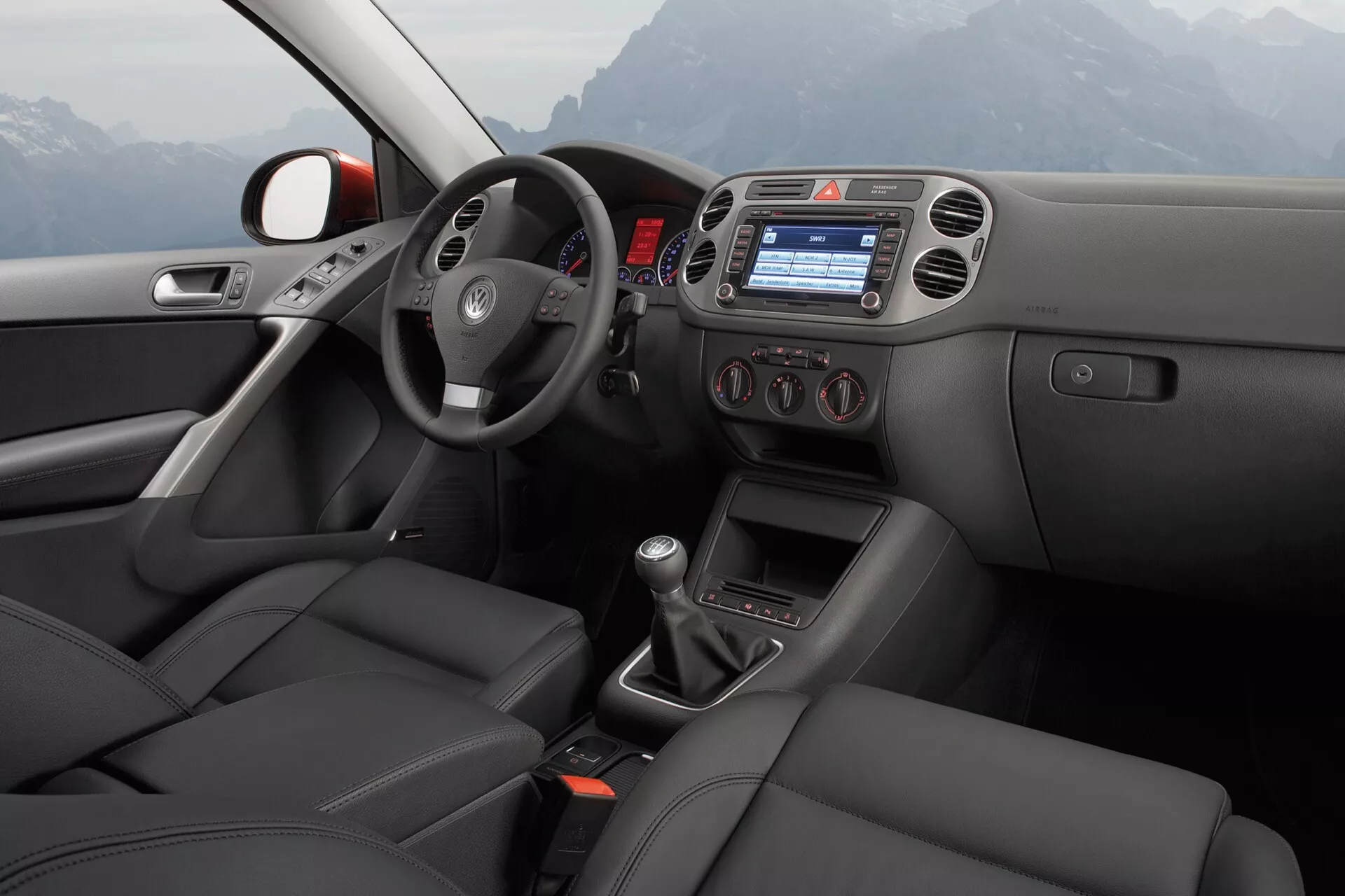 Vue intérieure du VW Tiguan 2007 depuis le point de vue du passager arrière, montrant la porte du conducteur avec commandes électriques pour les fenêtres et les rétroviseurs, volant avec commandes intégrées, console centrale avec écran d'infodivertissement intégré, commande climatique, sélecteur de vitesse manuel, sièges style sport en cuir, en état impeccable