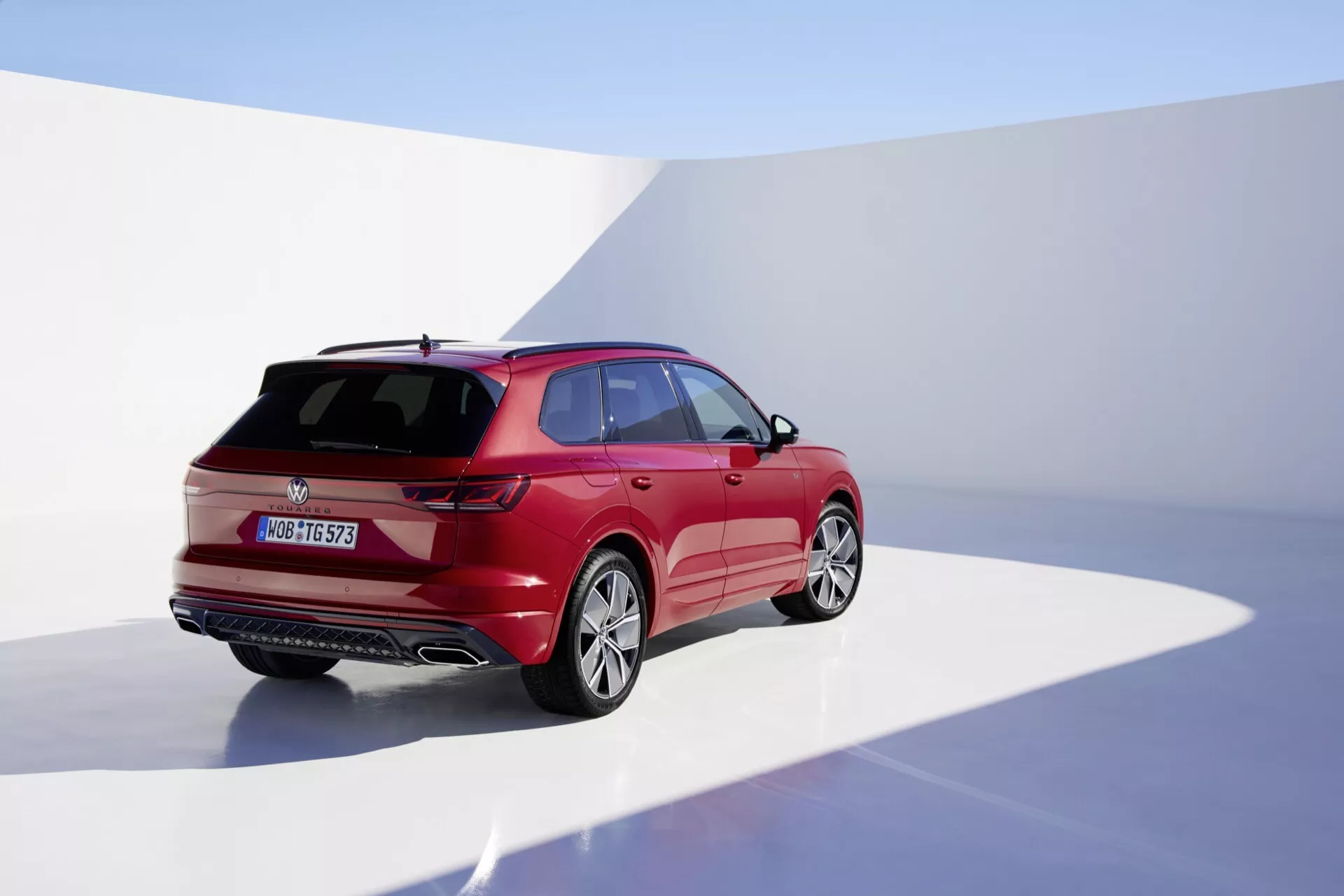 Modèle 2023 VW Touareg III facelift photographié depuis une vue arrière trois quarts mettant en lumière le côté droit, en studio avec fond continu, exhibant sa finition rouge brillante, design aérodynamique avec des lignes de carrosserie nettes, feux arrière LED, essuie-glace arrière, spoiler monté sur le toit et roues alliage.