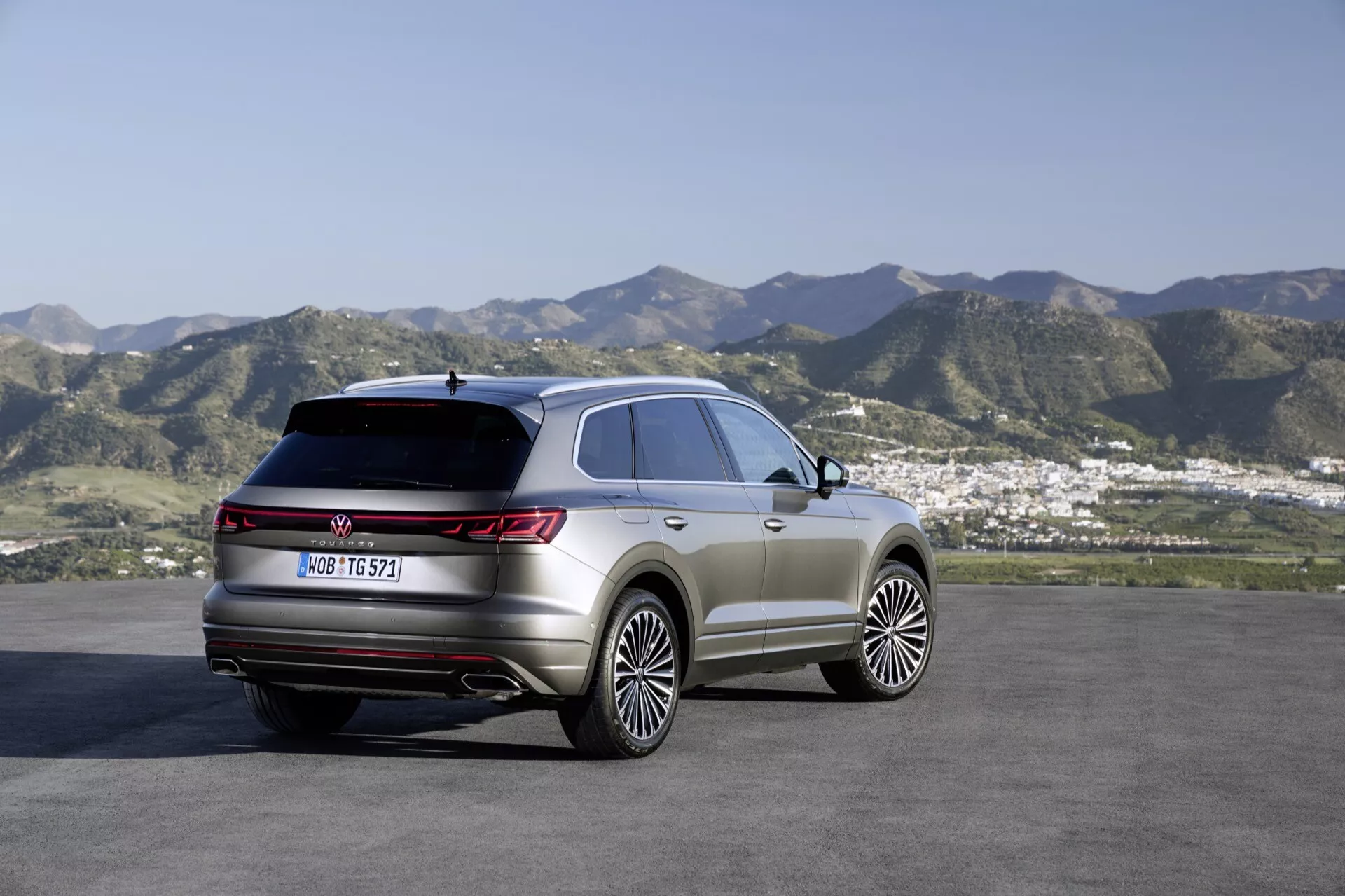 SUV VW Touareg 2023 (Facelift III) garé en extérieur avec en arrière-plan un paysage vallonné et une petite ville ou zone suburbaine, montrant le quart arrière droit et le profil latéral, mettant en évidence les feux arrière, la ligne de toit et les lignes de contour du corps. Caractéristiques comprennent de grandes roues en alliage, toit panoramique, feux arrière LED et éléments de style off-road premium.