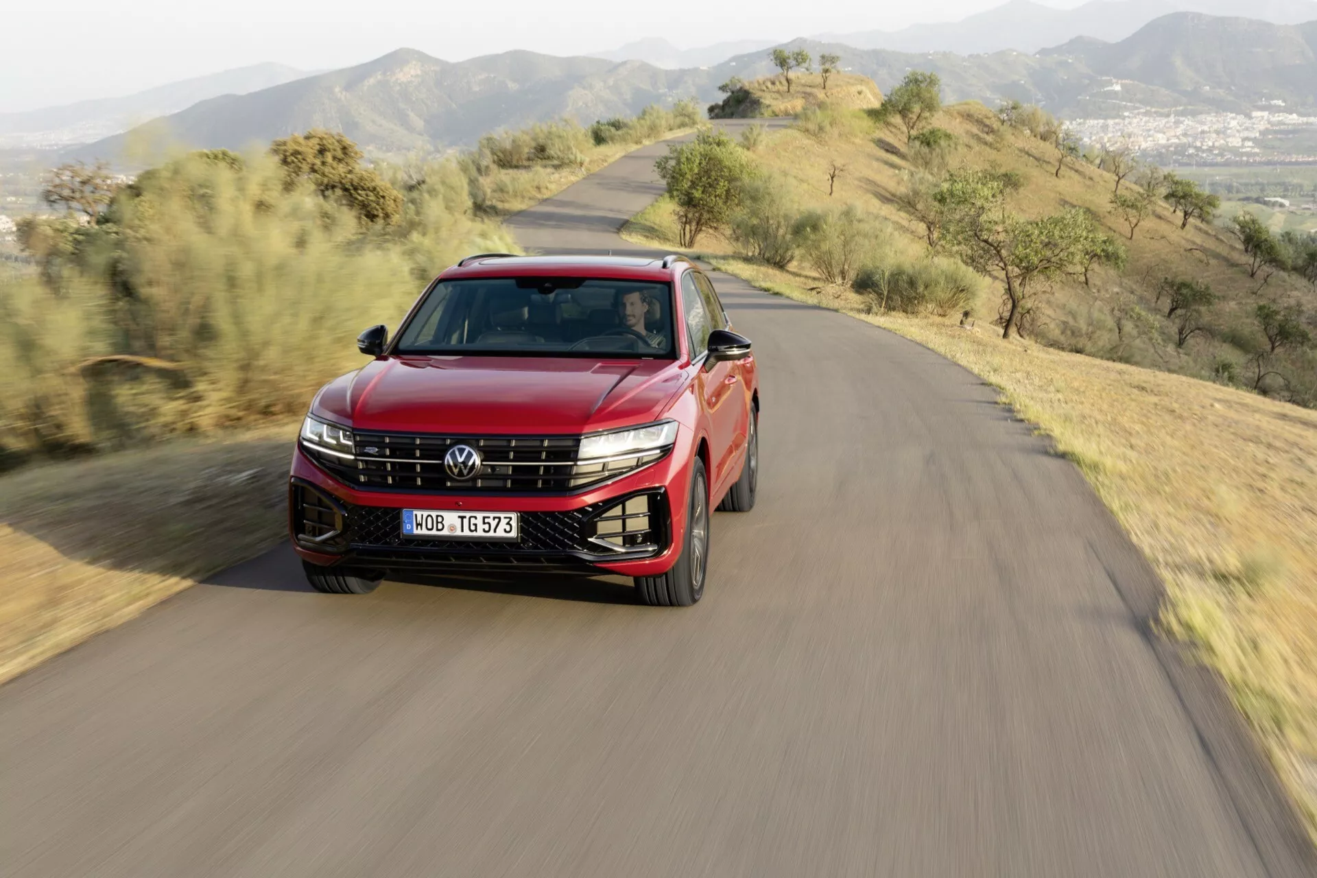 VW Touareg 2023 (III Facelift) crossover/SUV conduisant sur une route sinueuse de colline, avec phares LED, jantes en alliage, barres de toit, toit panoramique, peinture bicolore et rétroviseurs latéraux avec clignotants, capturé d’une perspective aérienne frontal et profil en plein jour