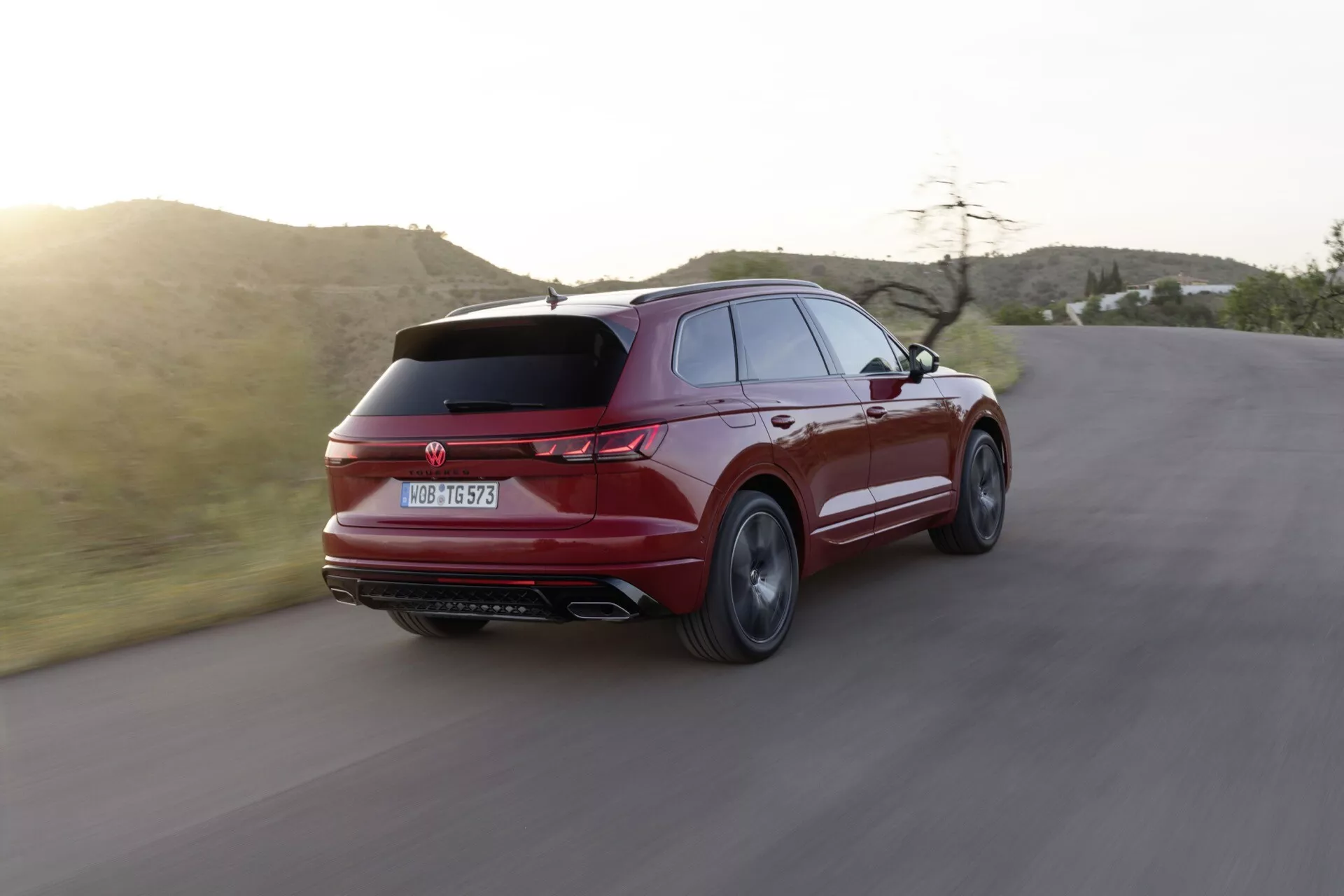 Facelift du Volkswagen Touareg III 2023 capturé dans une vue arrière dynamique de trois quarts sur une route sinueuse, mettant en évidence son design sportif et élégant, son spoiler arrière élégant, ses feux arrière distinctifs, ses doubles tuyaux d'échappement et ses roues en alliage à rayons multiples se détachant sur un paysage semi-aride à l'heure dorée