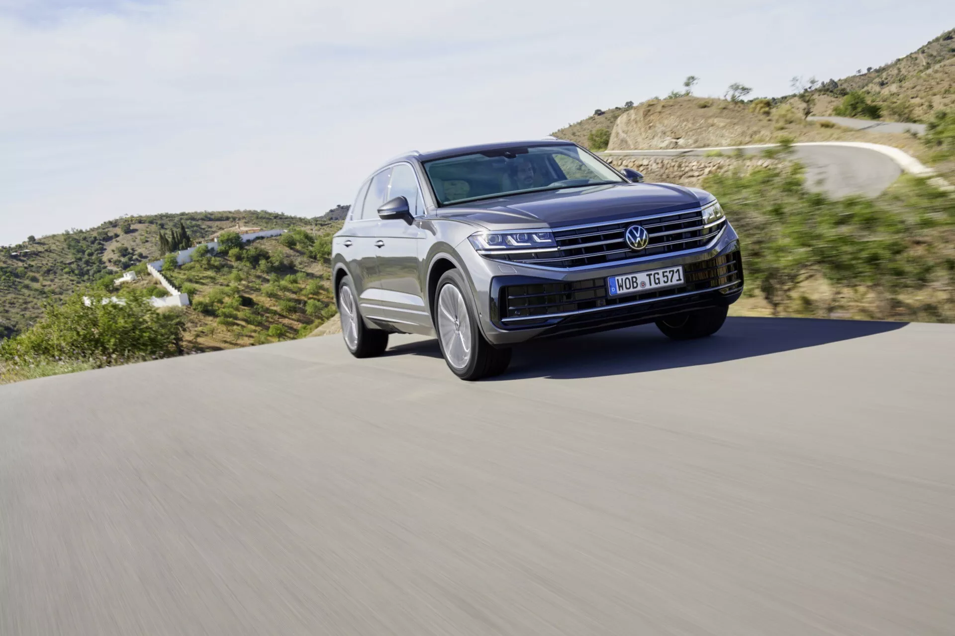 SUV Volkswagen Touareg III Facelift de 2023 en mouvement sur une route asphaltée, présentant une grille avant proéminente, des phares LED et le contour élégant du capot sur fond de paysage naturel, soulignant le design moderne et la technologie automobile.