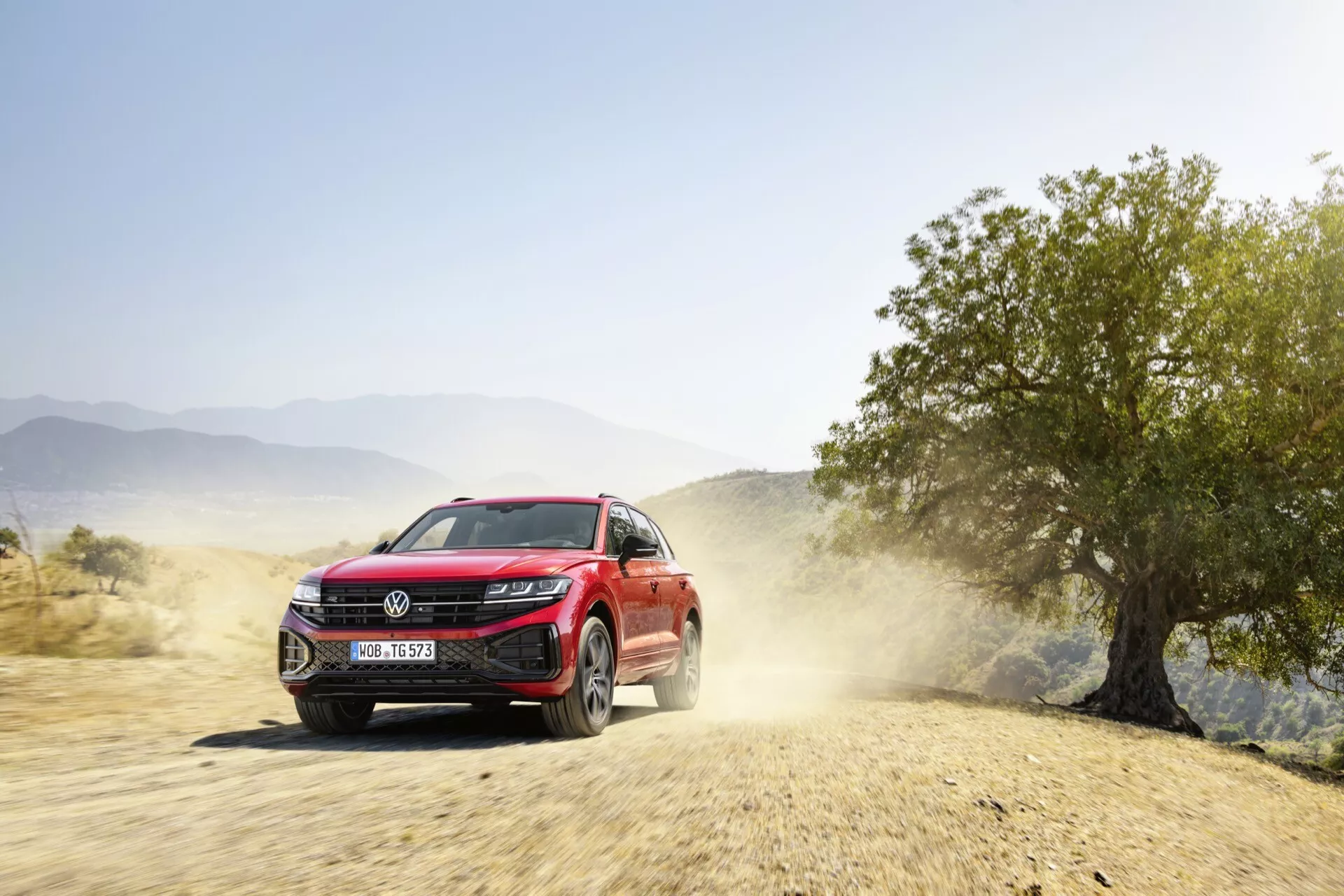 SUV rouge VW Touareg 2023 en mouvement dynamique sur une route non pavée avec un sillage de poussière, montrant la façade avant, les phares LED, la grille avec de potentiels capteurs d'assistance au conducteur et les roues en alliage, indiquant des capacités tout-terrain et des caractéristiques technologiques modernes