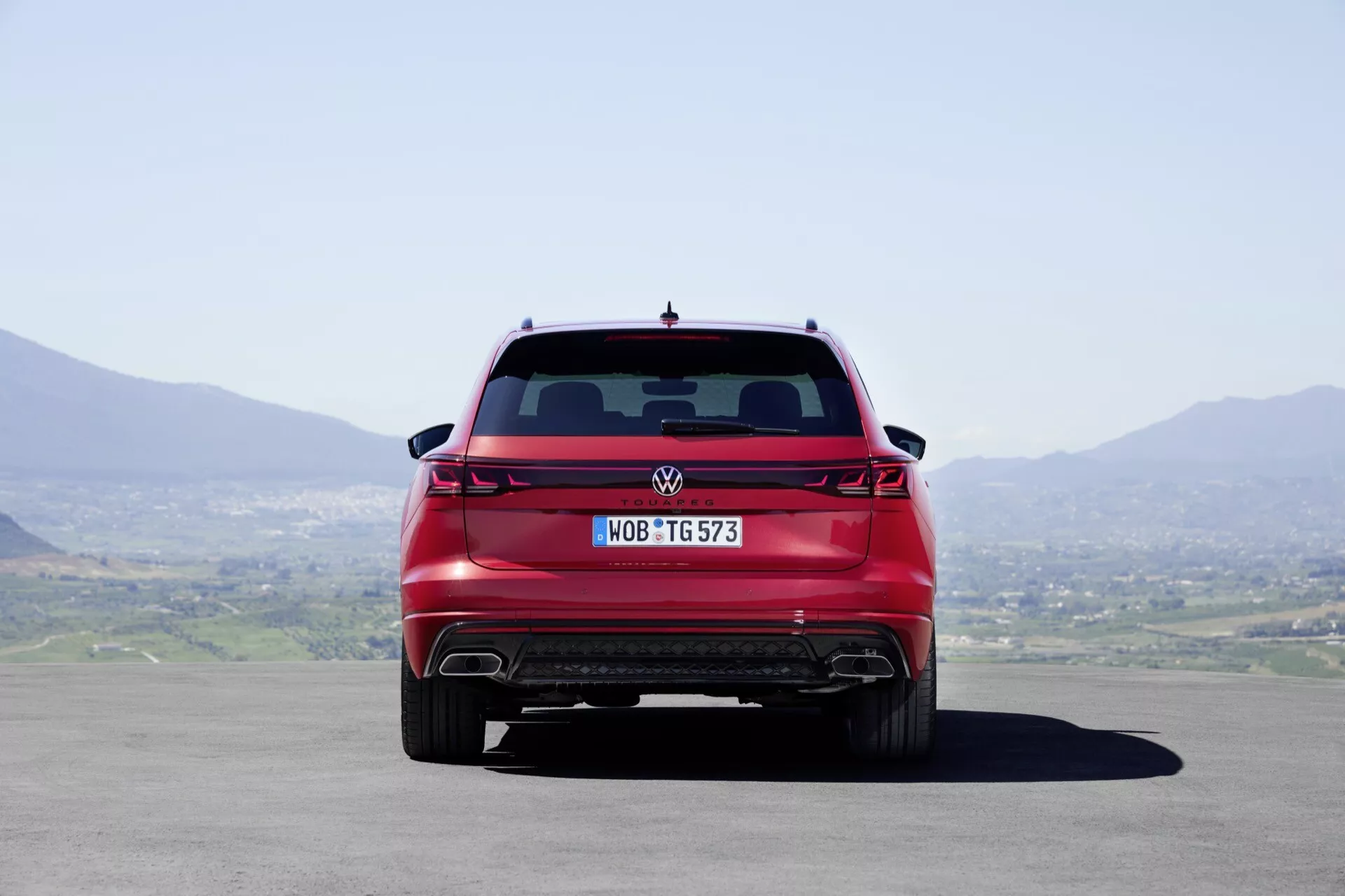 Vue arrière du Volkswagen Touareg 2023 montrant la largeur complète, feux arrière modernes, diffuseur arrière et embouts d'échappement doubles, sous la lumière naturelle du jour avec un ciel clair et un paysage vallonné en arrière-plan