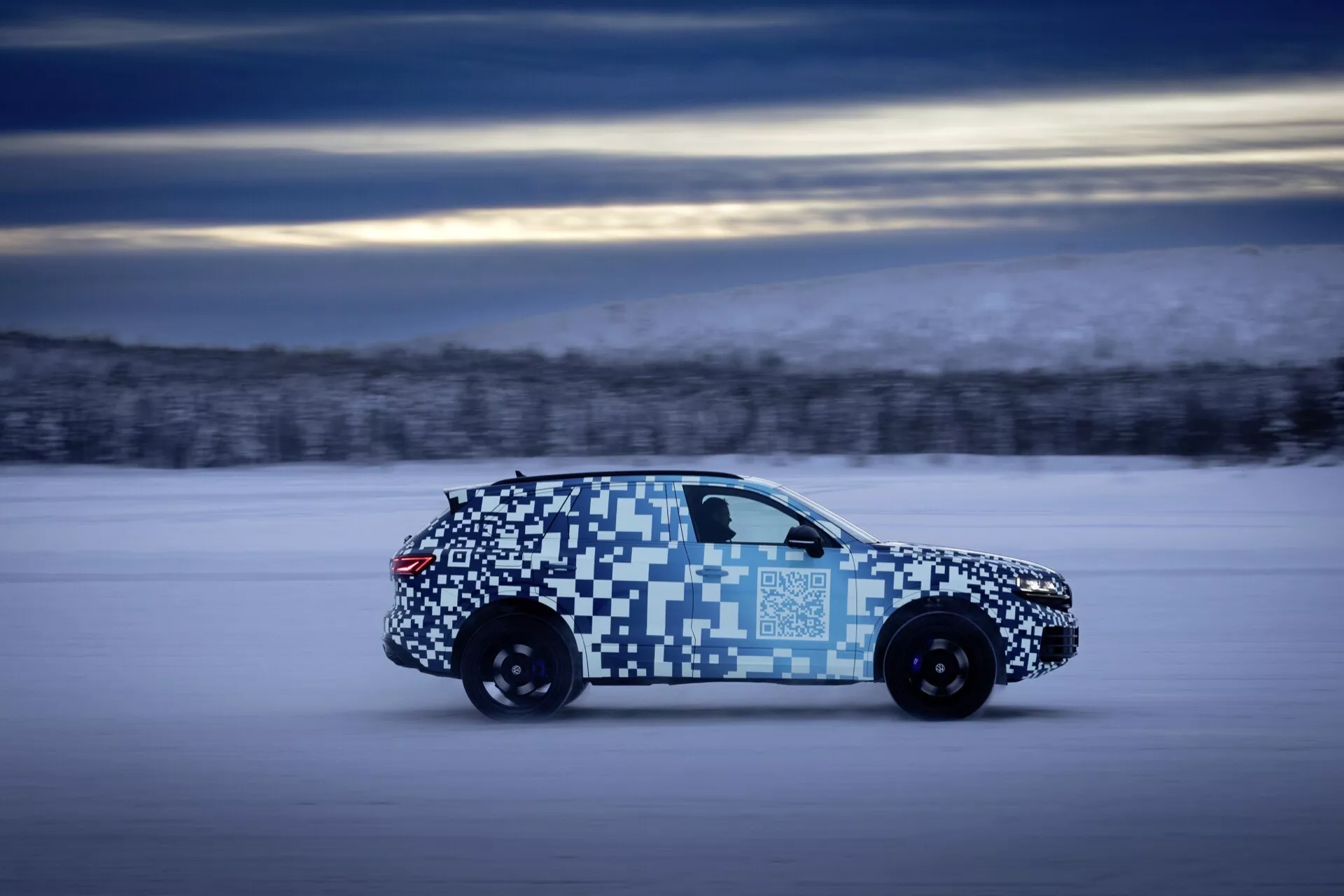 VW Touareg 2023 (III-restylage) en mouvement, soulignant son design aérodynamique et ses feux arrière LED, avec un emballage de camouflage, dans un cadre de test hivernal