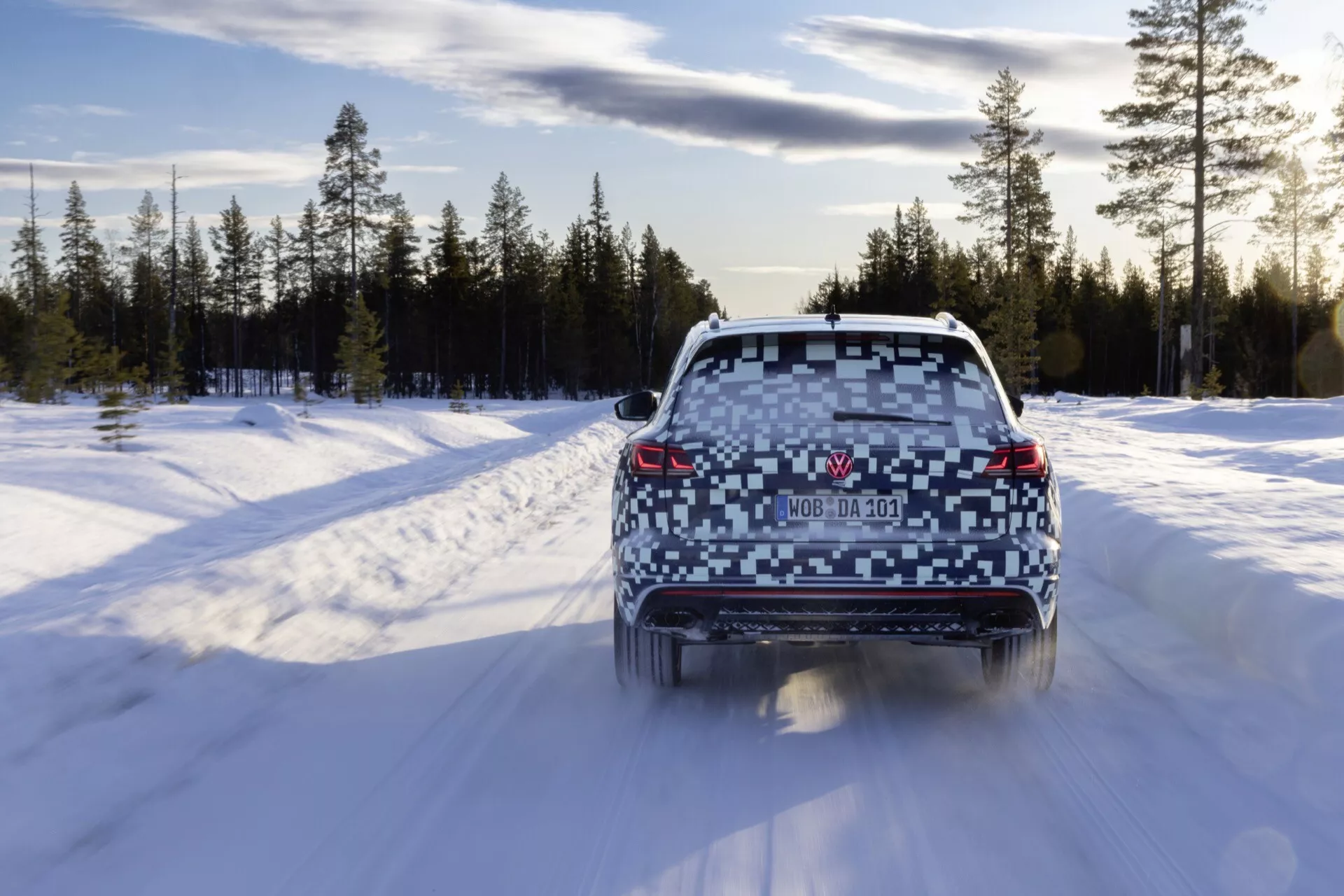 Vue arrière du Volkswagen Touareg 2023 (III-Facelift) sur une route enneigée dans un paysage forestier hivernal, montrant des feux arrière allumés, un pare-brise arrière clair, des sorties d'échappement doubles et un aileron de toit.