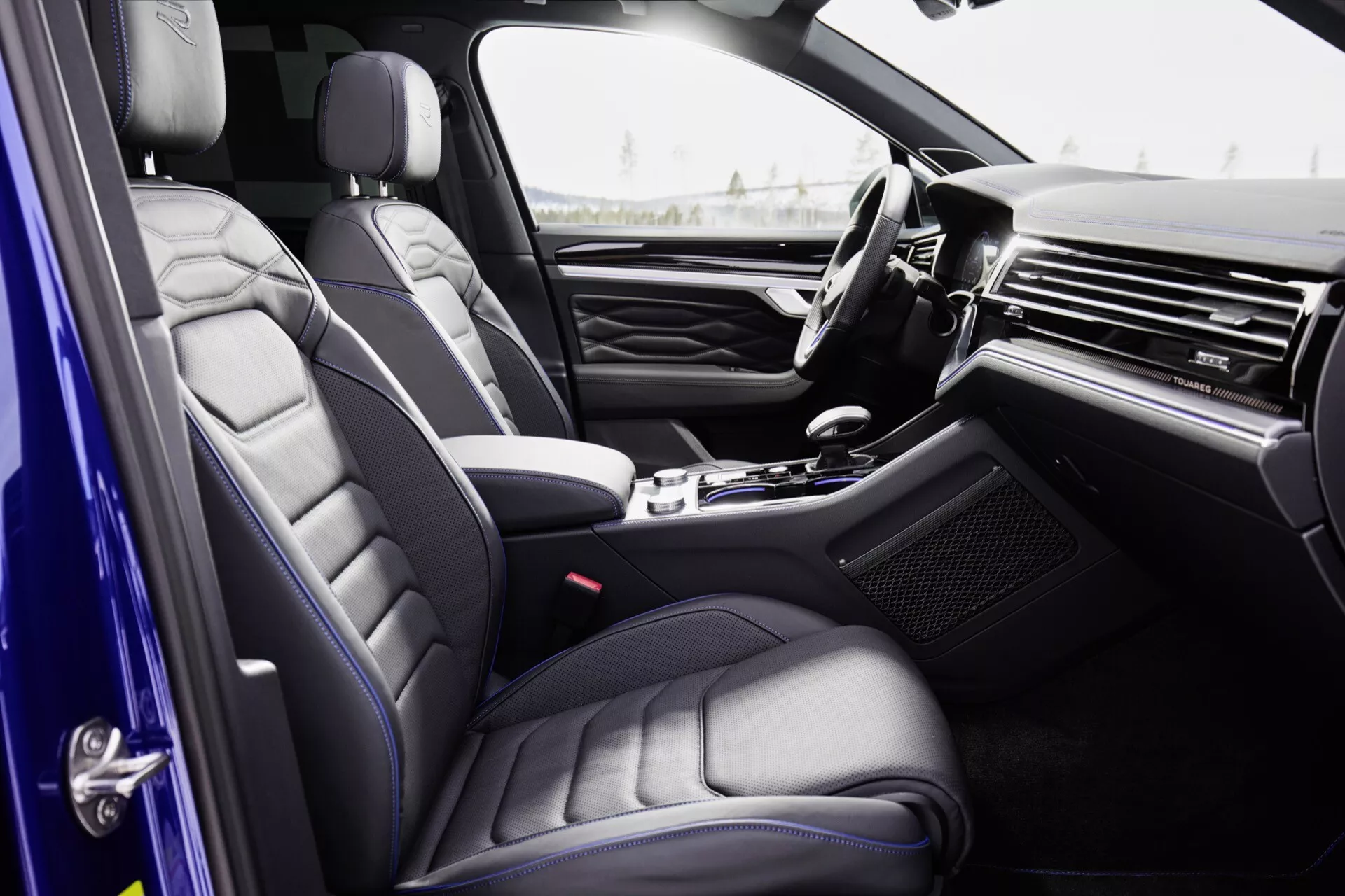Vue intérieure du Volkswagen Touareg 2023 (III facelift) depuis la perspective du passager, mettant en avant des sièges en cuir haut de gamme avec un design matelassé, un tableau de bord élégant avec des garnitures métalliques ou en bois, une console centrale de luxe avec sélecteur de vitesse automatique, un panneau de porte à options élevées, et un volant partiellement visible avec des commandes intégrées.