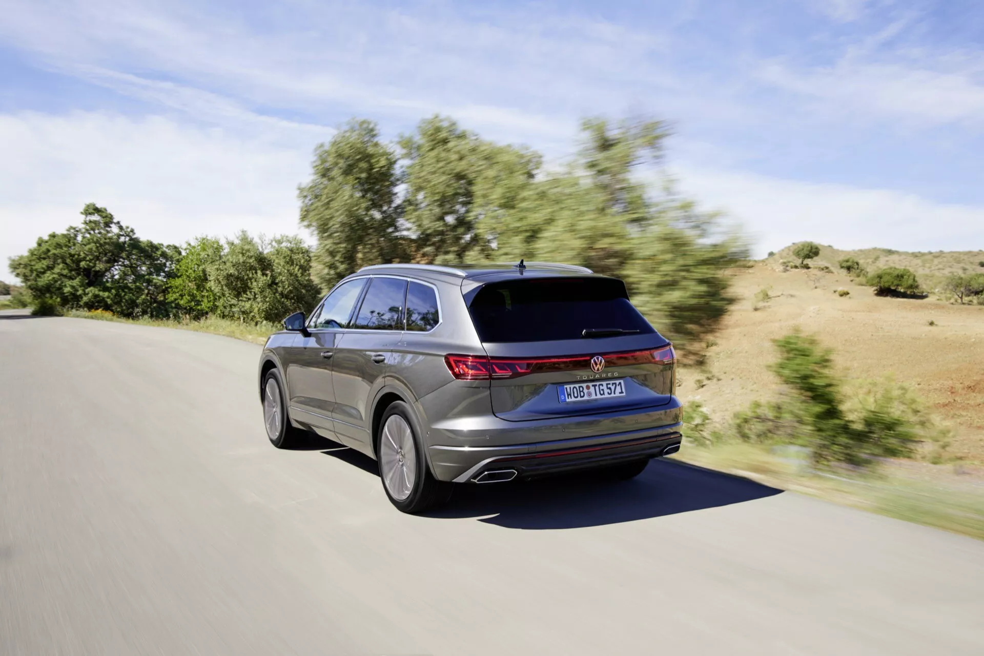 VW Touareg III facelift 2023 capturé d’un angle arrière trois-quarts en mouvement pendant la journée par temps clair, montrant des feux arrière LED, un design élégant de break avec une ligne de toit inclinée et un becquet de toit, un aperçu partiel du design de la jante alliage, et une finition de peinture métallique.
