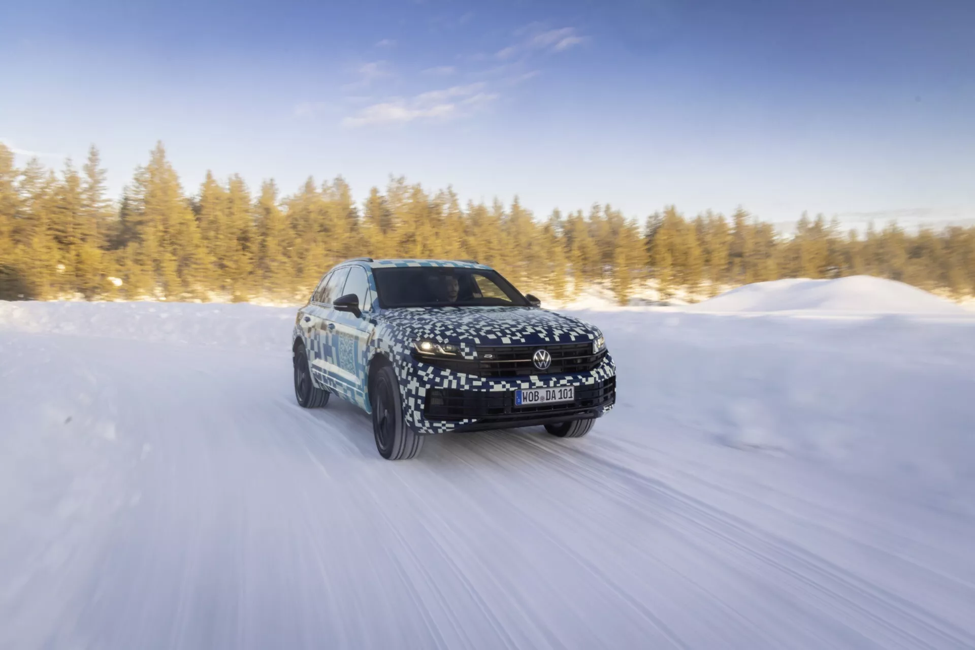 Modèle Volkswagen Touareg III facelift 2023 conduisant dynamiquement sur route enneigée sous le soleil, montrant un mouvement vers l'avant, phares LED, roues en alliage et son design aérodynamique avec enveloppe de camouflage.