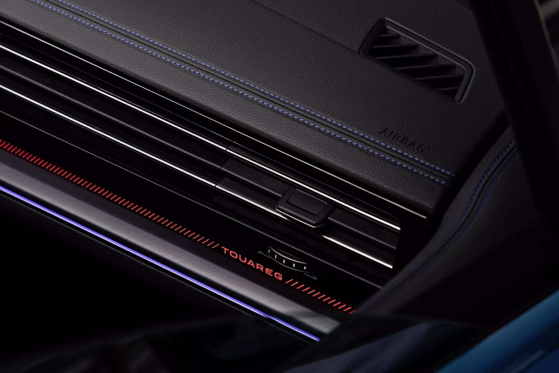 Vue rapprochée de l'intérieur du Volkswagen Touareg facelift 2023 mettant en vedette le design du tableau de bord, notamment l'inscription 'Touareg' avec éclairage ambiant, des bouches d'aération avec accents argentés et une barre lumineuse rouge atmosphérique pour une sensation de luxe