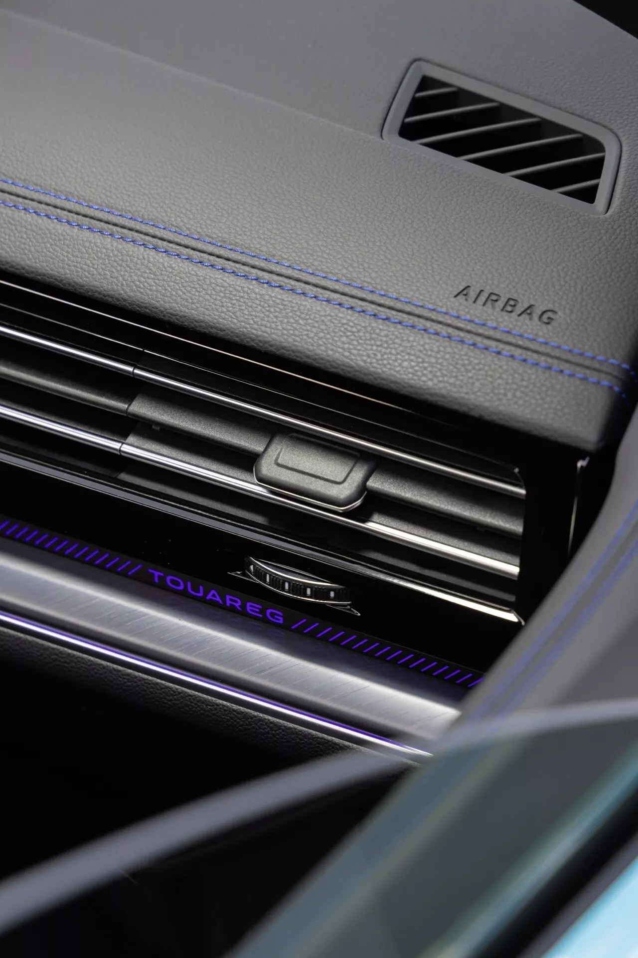 Vue rapprochée et angulaire du tableau de bord côté passager de la Volkswagen Touareg 2023 (III-Facelift) soulignant l'indication AIRBAG, les lamelles de ventilation de la climatisation finies en chrome avec un bouton de contrôle central, une plaque nominative 'TOUAREG' illuminée, des matériaux luxueux avec coutures contrastées, et des éléments de design intérieur raffinés dans un environnement à faible luminosité mettant en évidence la technologie d'éclairage intérieur avancée du véhicule et les caractéristiques de sécurité.