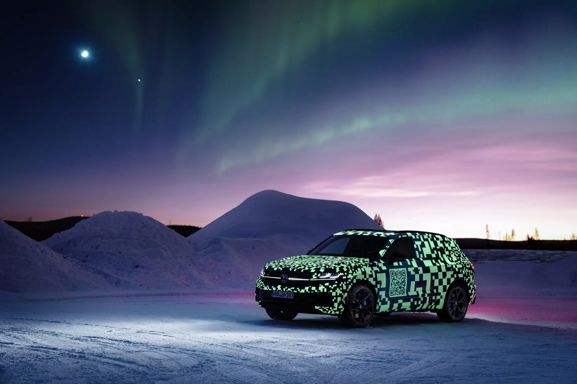 Prototype VW Touareg (III-facelift) en pellicule de camouflage sur surface enneigée sous les aurores boréales avec un astre brillant dans le ciel crépusculaire, mettant en avant l'éclairage avancé et la technologie électrique ou hybride potentielle.