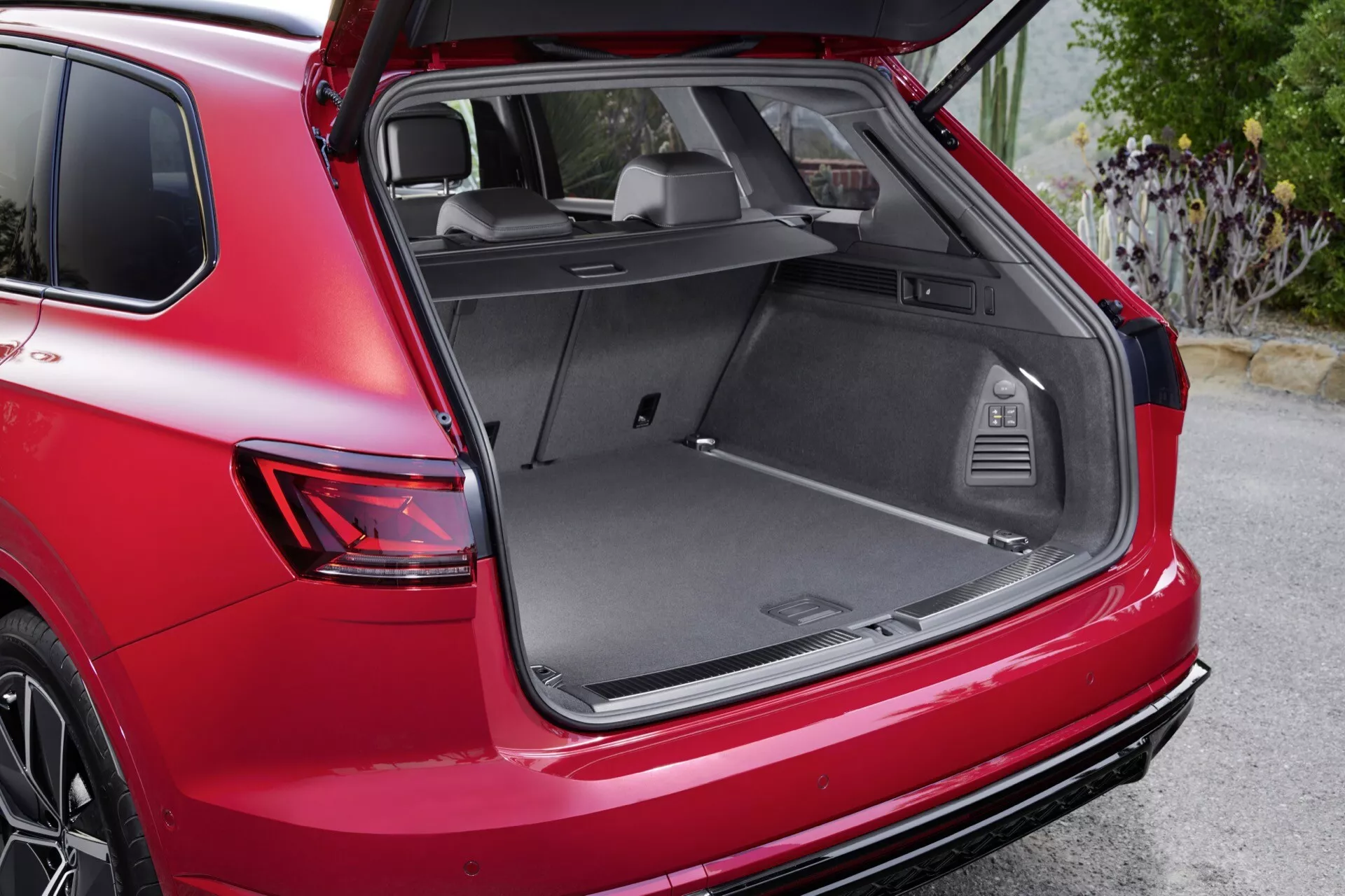 SUV rouge VW Touareg III facelift 2023 vue arrière trois quarts avec coffre ouvert sur surface pavée montrant l'espace de chargement, le design des feux arrière et la roue arrière