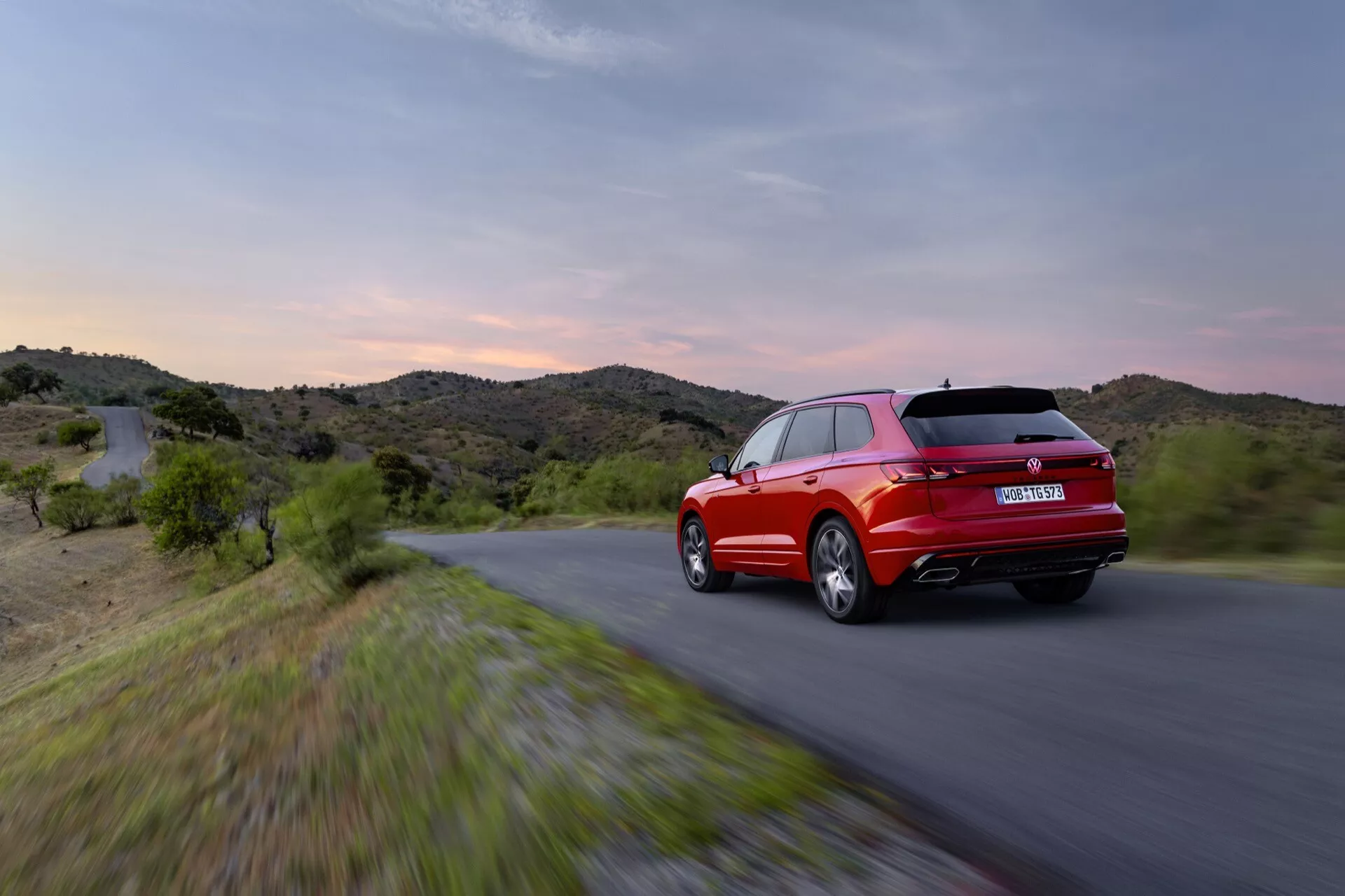 VW Touareg III Facelift 2023 en mouvement vu de trois quarts arrière avec feux arrière LED et contour sportif sur fond de ciel crépusculaire et de collines, mettant en avant sa couleur rouge, son design aérodynamique et ses caractéristiques de performance.
