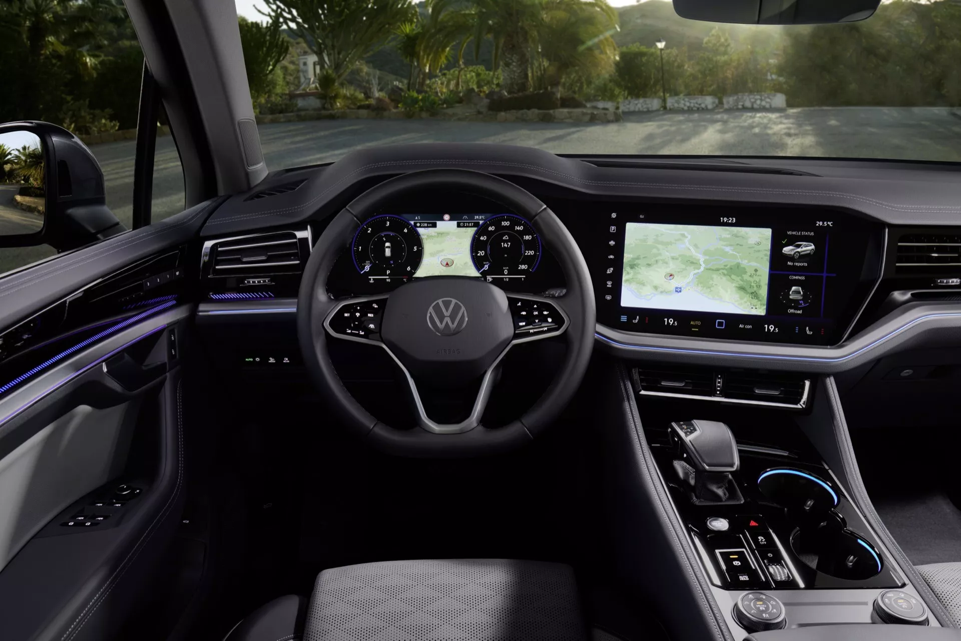 Vue intérieure du Volkswagen Touareg III facelift 2023 depuis les sièges arrières vers le tableau de bord numérique éclairé par la lumière naturelle, mettant en avant le volant multifonctions avec logo, le combiné d'instruments numérique, le grand système d'infodivertissement tactile avec GPS, le contrôle climatique tactile élégant, le levier de vitesses automatique minimaliste, et la console centrale finie de haute qualité avec éclairage d'ambiance et fonctionnalités de sécurité