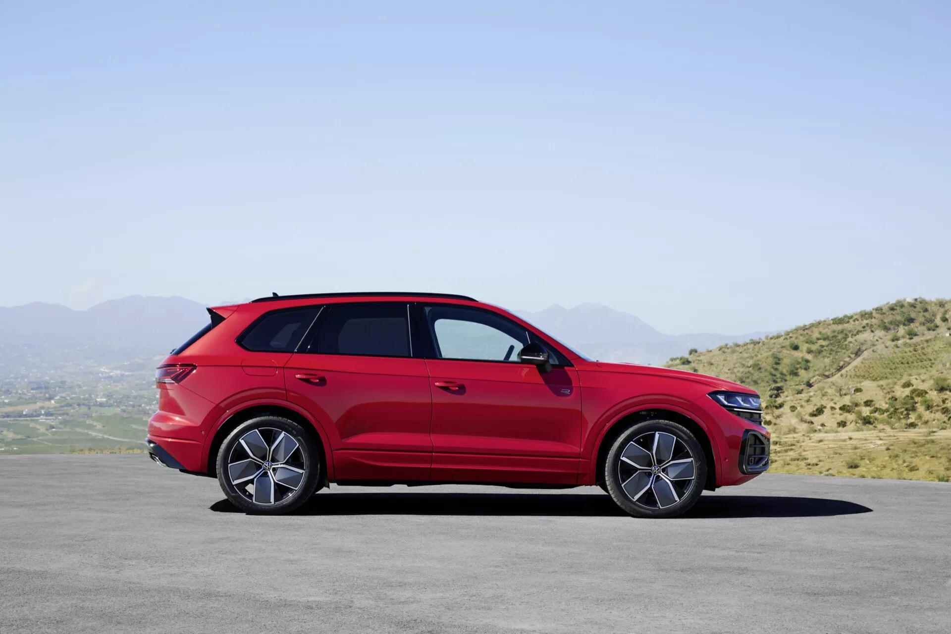 Modèle VW Touareg III lifting 2023 en rouge stationné sur une surface plate avec un paysage montagneux et un ciel clair en arrière-plan, montrant son profil latéral avec une vue détaillée des roues en alliage, des poignées de portes, des rétroviseurs latéraux avec clignotants intégrés, des barres de toit, une posture sportive style coupé et des vitres teintées.