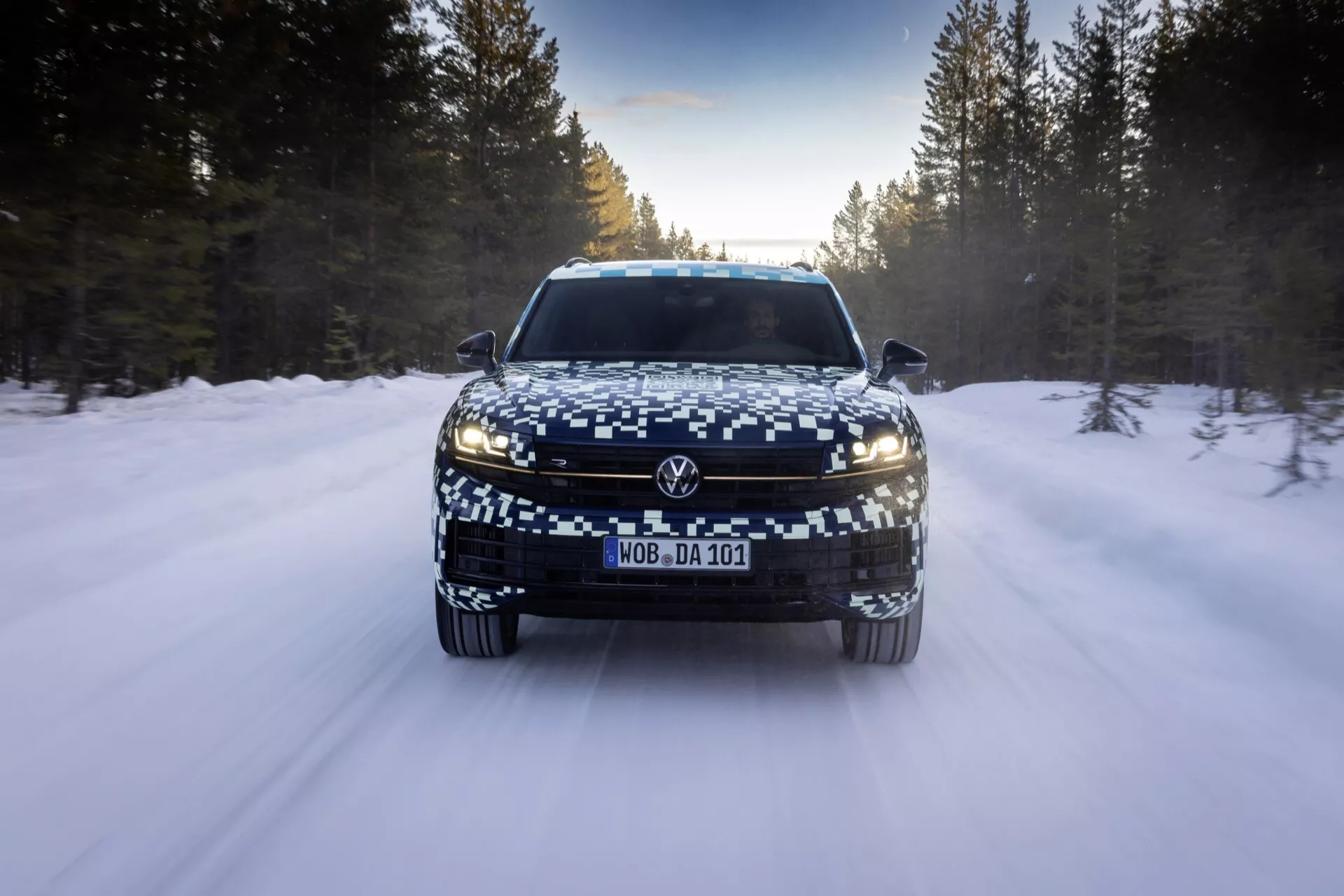 Vue de face du VW Touareg 2023 (III-Facelift) sur une route enneigée, montrant des phares LED, une large grille et une plaque d'immatriculation camouflée, reflétant des fonctionnalités éclairantes avancées et des capacités de conduite hivernale.