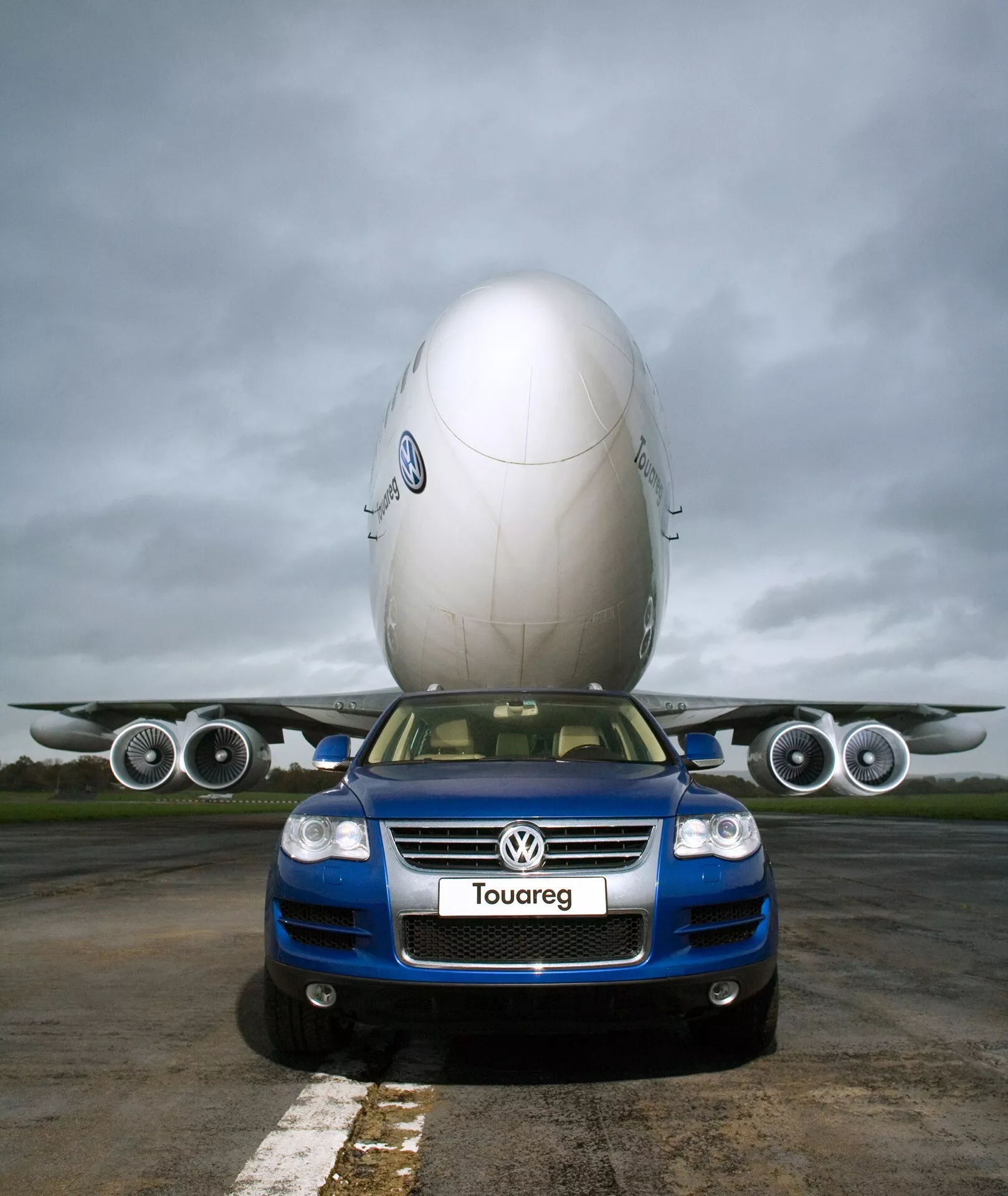Volkswagen Touareg 2023 (III-Facelift) bleu, positionnée en premier plan sur le tarmac d'un aéroport, capturée sous un angle bas soulignant sa taille et sa facade avant, dotée d'une grille avec logo, de phares, de feux de brouillard et d'un badge Touareg, avec un arrière-plan créatif d'un jet commercial semblant reposer sur son toit contre un ciel nuageux, mettant en valeur la conception robuste et les caractéristiques technologiques du véhicule