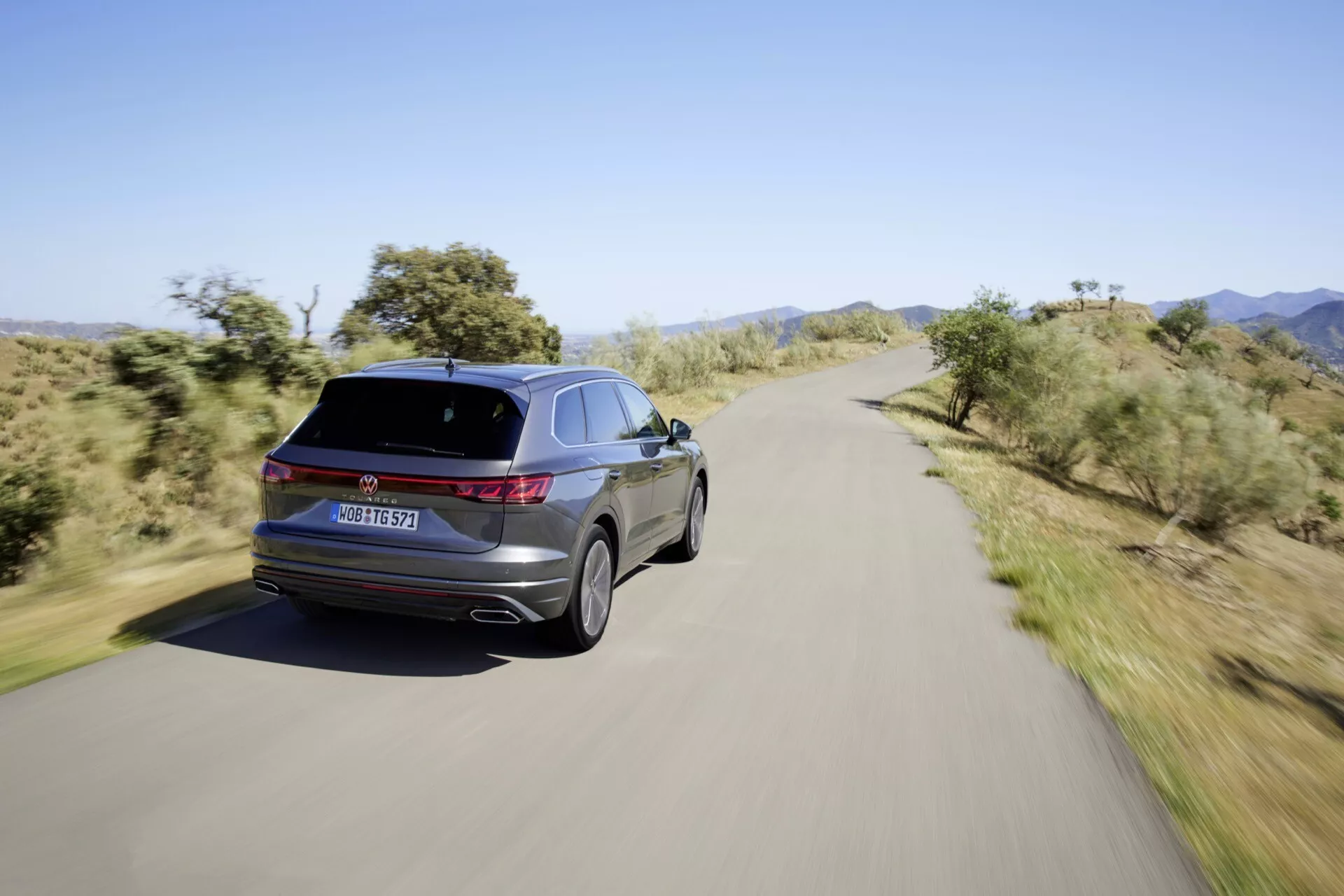 Vue arrière trois-quarts de la VW Touareg III-Facelift 2023 sur une route pittoresque, affichant des feux arrière LED, des badges distinctifs, des embouts d'échappement doubles, des jantes en alliage, une ligne de toit élégante et une finition brillante.
