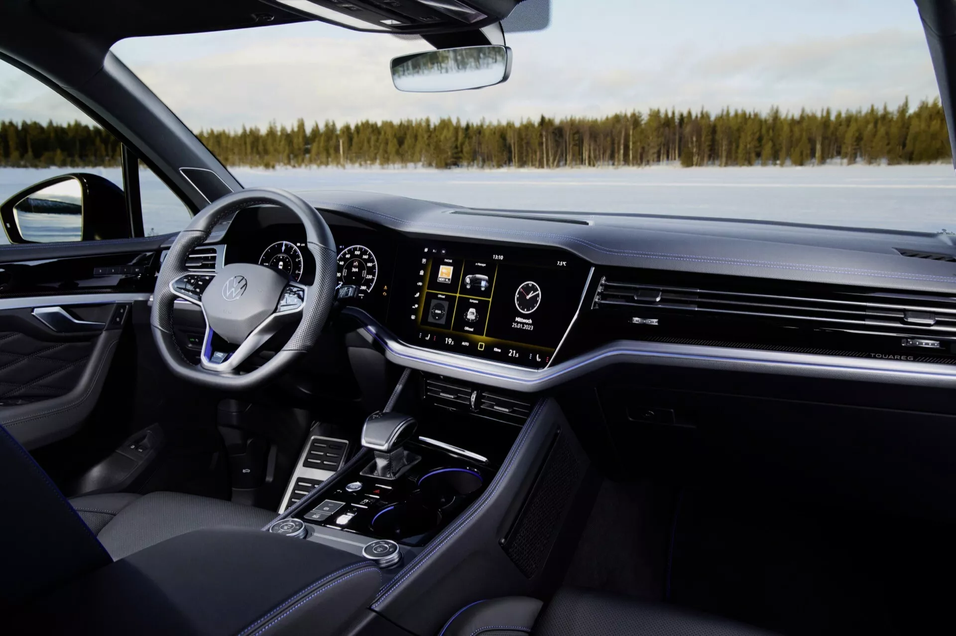Vue intérieure du VW Touareg 2023 depuis le siège passager avant présentant un siège en cuir noir, une console centrale avec levier de vitesse, porte-gobelets, boutons de contrôle, système d'infodivertissement tactile, bouches de climatisation, tableau de bord numérique, et volant avec contrôles, sur fond de paysage enneigé à travers le pare-brise, mettant en évidence les caractéristiques modernes de luxe et de technologie du véhicule.