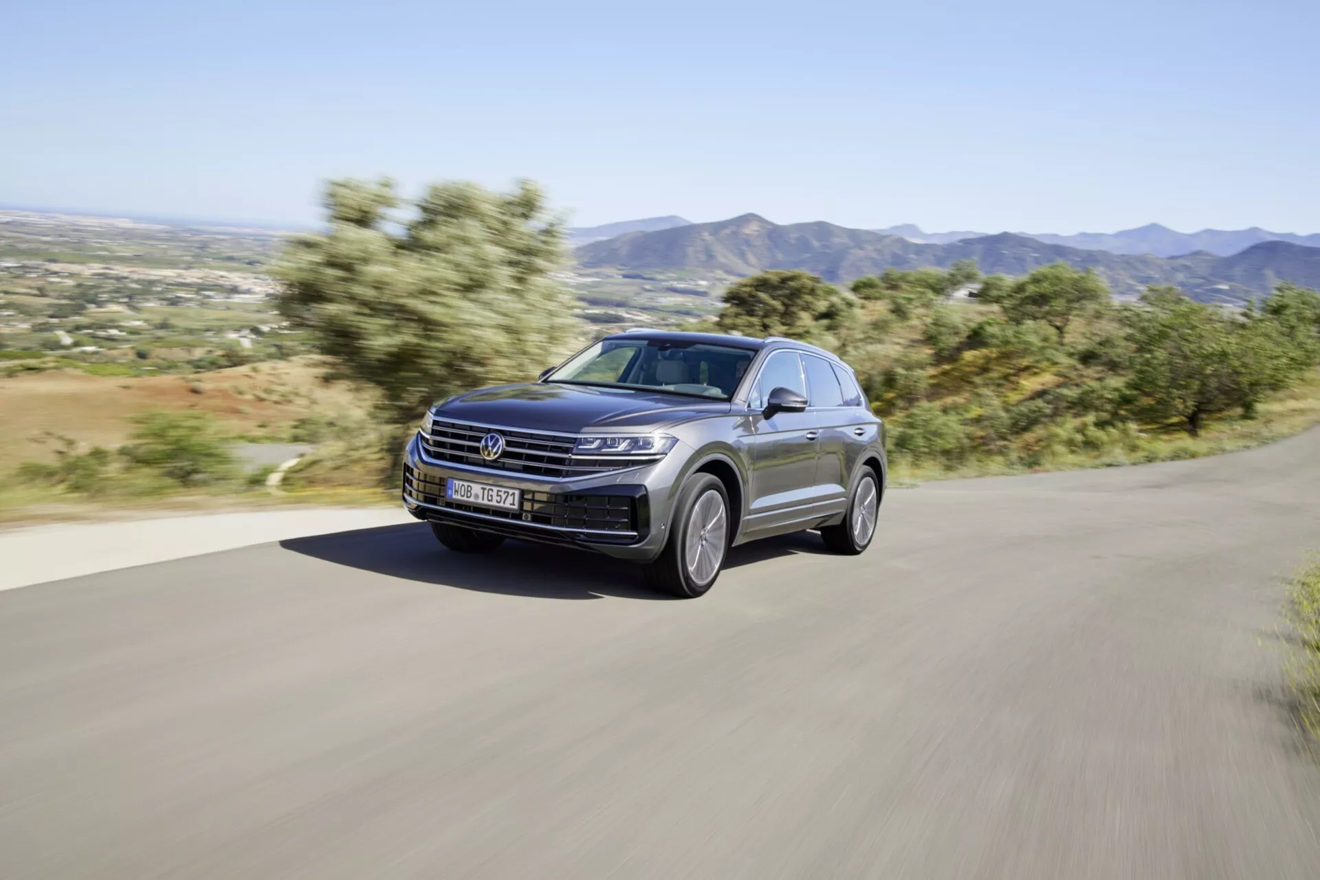 VW Touareg 2023 facelift roulant dynamiquement sur une route rurale pavée, présentant son design moderne et ses caractéristiques de performance, y compris les phares LED, la peinture métallisée de luxe et les roues en alliage élégantes, sur un fond montagneux