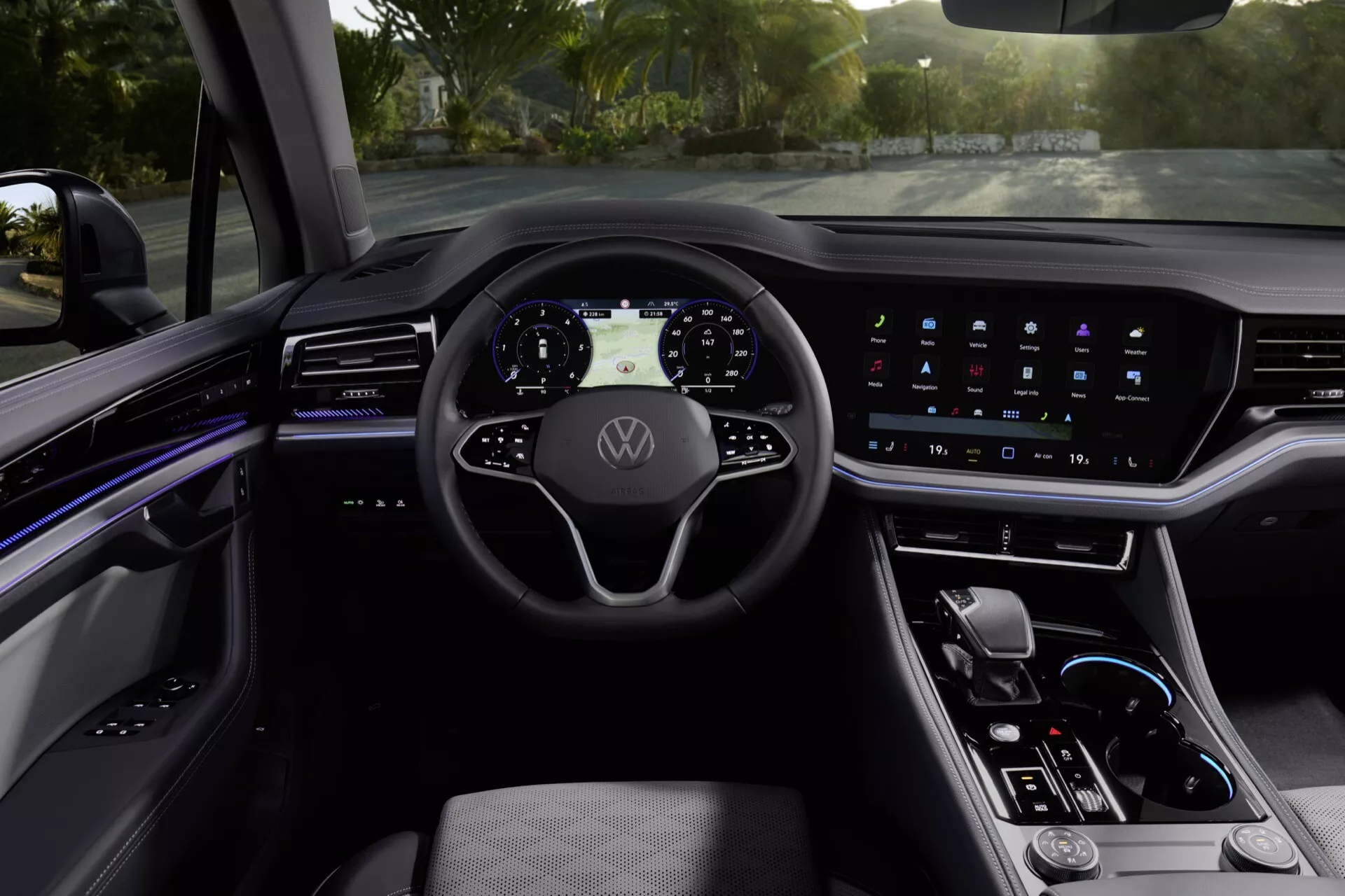 Vue intérieure du VW Touareg 2023 depuis la perspective du conducteur montrant le volant moderne à trois branches avec le logo Volkswagen, le tableau de bord numérique, le système d'infodivertissement à écran tactile large, l'éclairage d'ambiance, la console centrale élégante avec sélection de vitesse et matériaux haut de gamme