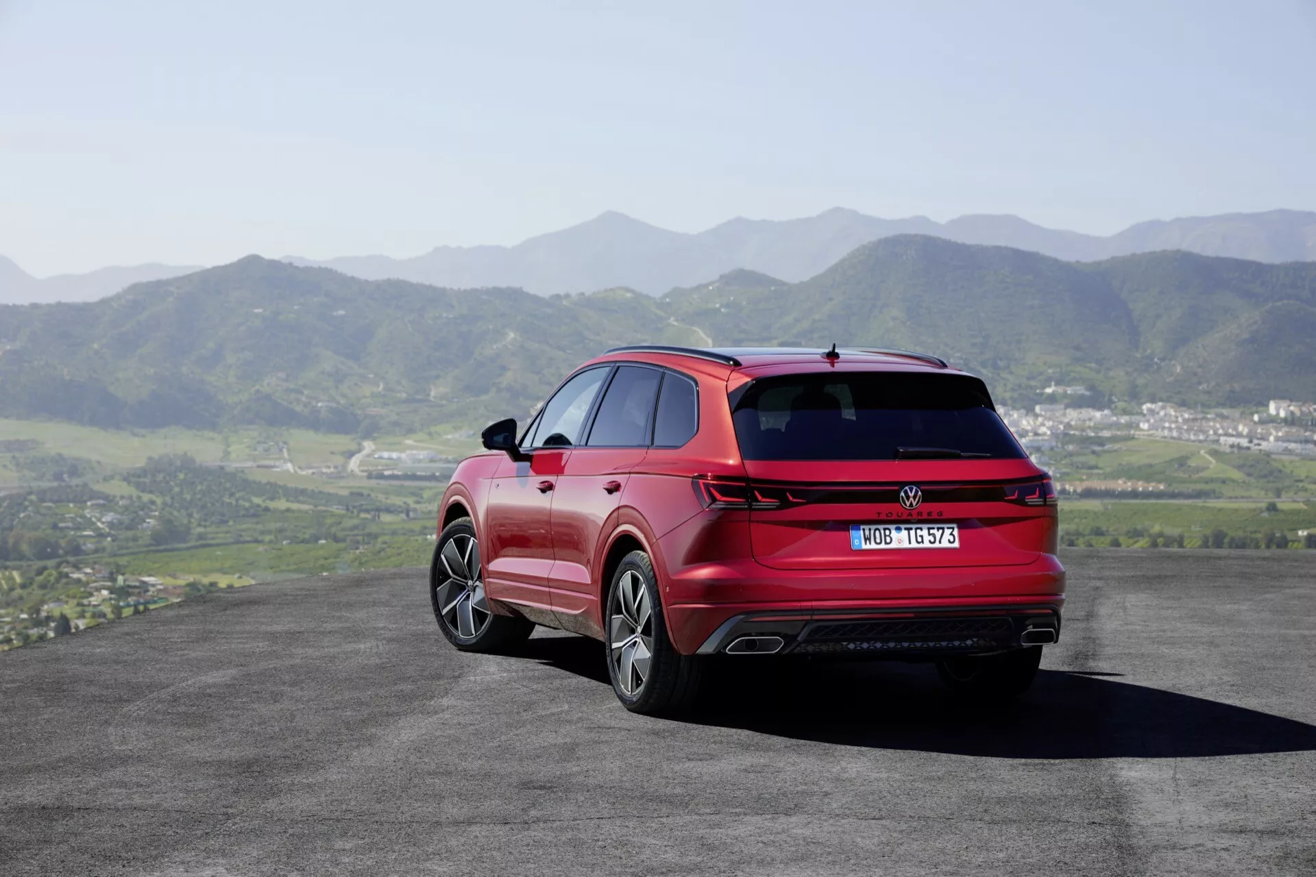 VW Touareg III Facelift 2023 en rouge - vue arrière et latérale montrant des feux arrière LED, des doubles sorties d'échappement, des jantes en alliage et des barres de toit argentées sur un fond de collines ondulantes et de ciel clair