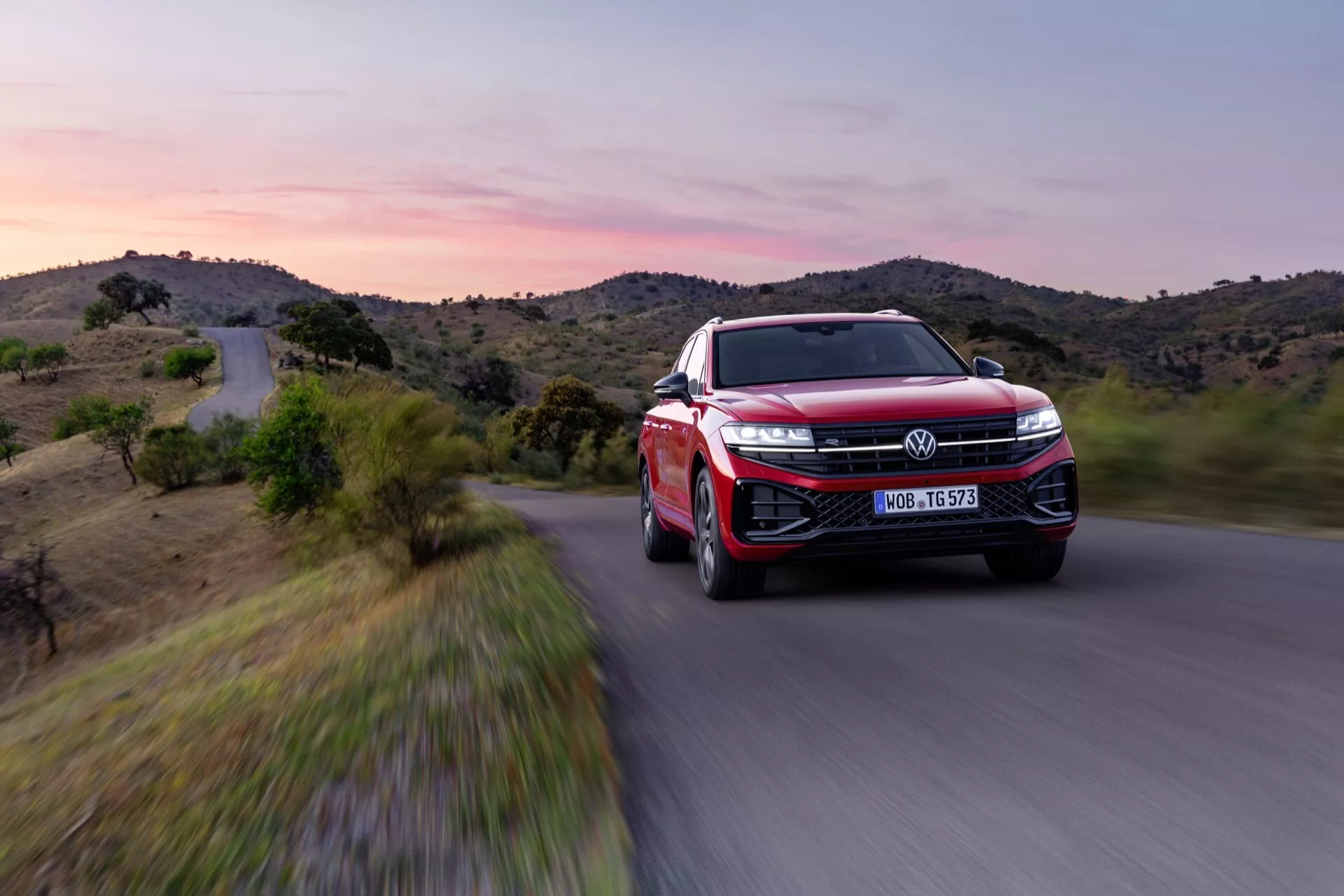 VW Touareg III-facelift 2023 en rouge, vue avant trois quarts mettant en avant les phares LED, grande grille avec emblème, design aérodynamique sportif, jantes en alliage avancées et caractéristiques ADAS en mouvement dynamique sur une route panoramique au crépuscule
