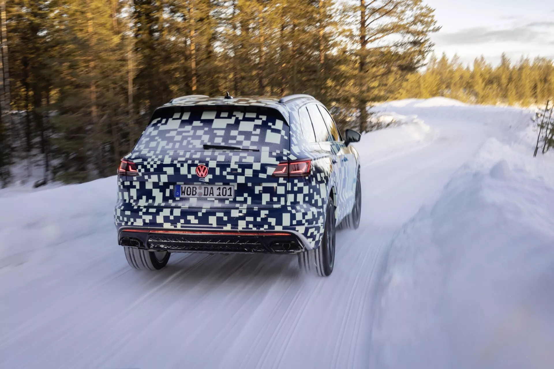 VW Touareg III Facelift 2023 en camouflage roulant sur une route couverte de neige, présentant des feux arrière LED, diffusor arrière, essuie-glace de fenêtre arrière, plaque d'immatriculation obscurcie, doubles embouts d'échappement, capteurs de pare-chocs arrière, flou de mouvement indiquant le mouvement, pneus d'hiver, et potentiel de traction intégrale