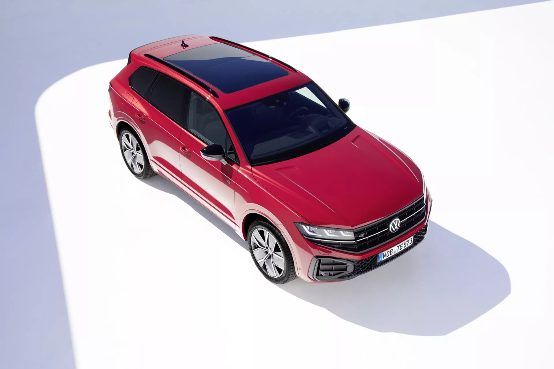 Vue en plongée du VW Touareg III Facelift 2023 de couleur rouge, présentant des caractéristiques de design telles que grille à lattes horizontales avec emblème, phares LED, pare-chocs avant aérodynamique avec prises d'air, lignes sculptées du capot, pare-brise expansif, jantes en alliage à multiples branches, toit panoramique et rétroviseurs latéraux aérodynamiques sur fond blanc