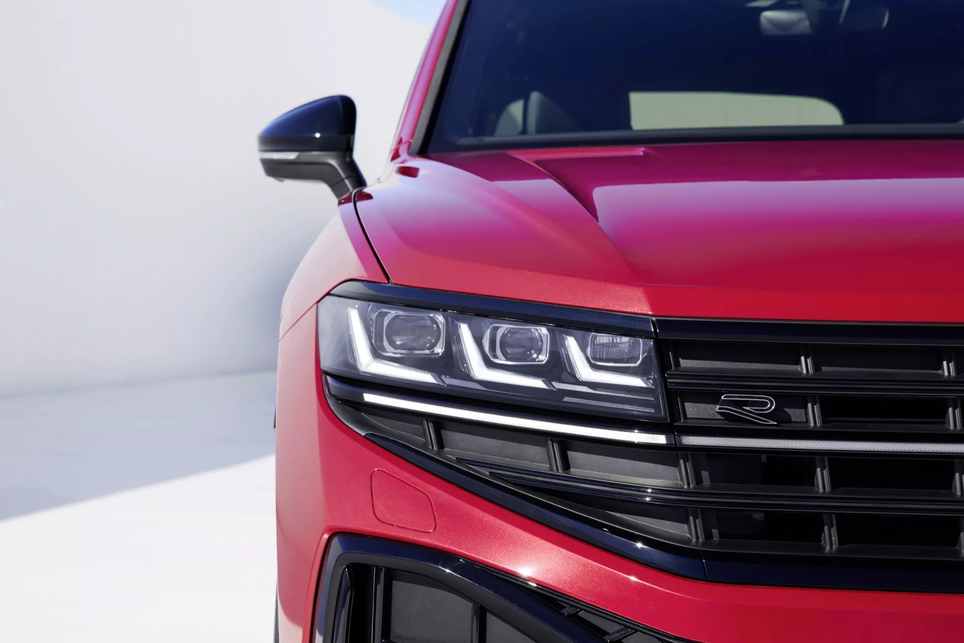 VW Touareg III-Facelift 2023 avec peinture rouge métallique brillant montrant une vue de près du quart avant gauche, montrant l'assemblage de phare LED, une partie de la grille à lamelles horizontales, une vue détaillée du pare-choc avant et des lignes aérodynamiques sur le capot, contre un fond flou soulignant le design moderne et les caractéristiques technologiques de la voiture.