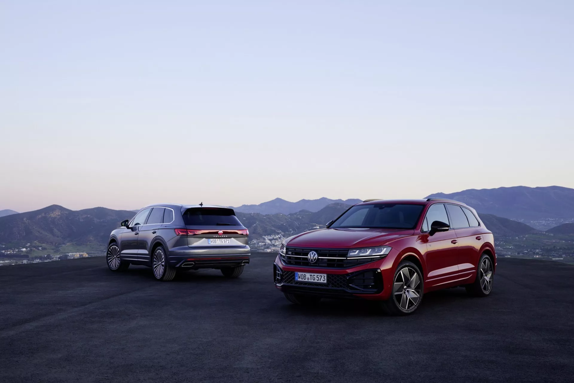 Modèles Volkswagen Touareg (III-facelift) 2023 en rouge et argent à l'extérieur avec un décor montagneux et un ciel crépusculaire, présentant des profils avant et latéral avec technologie d’éclairage LED avancée