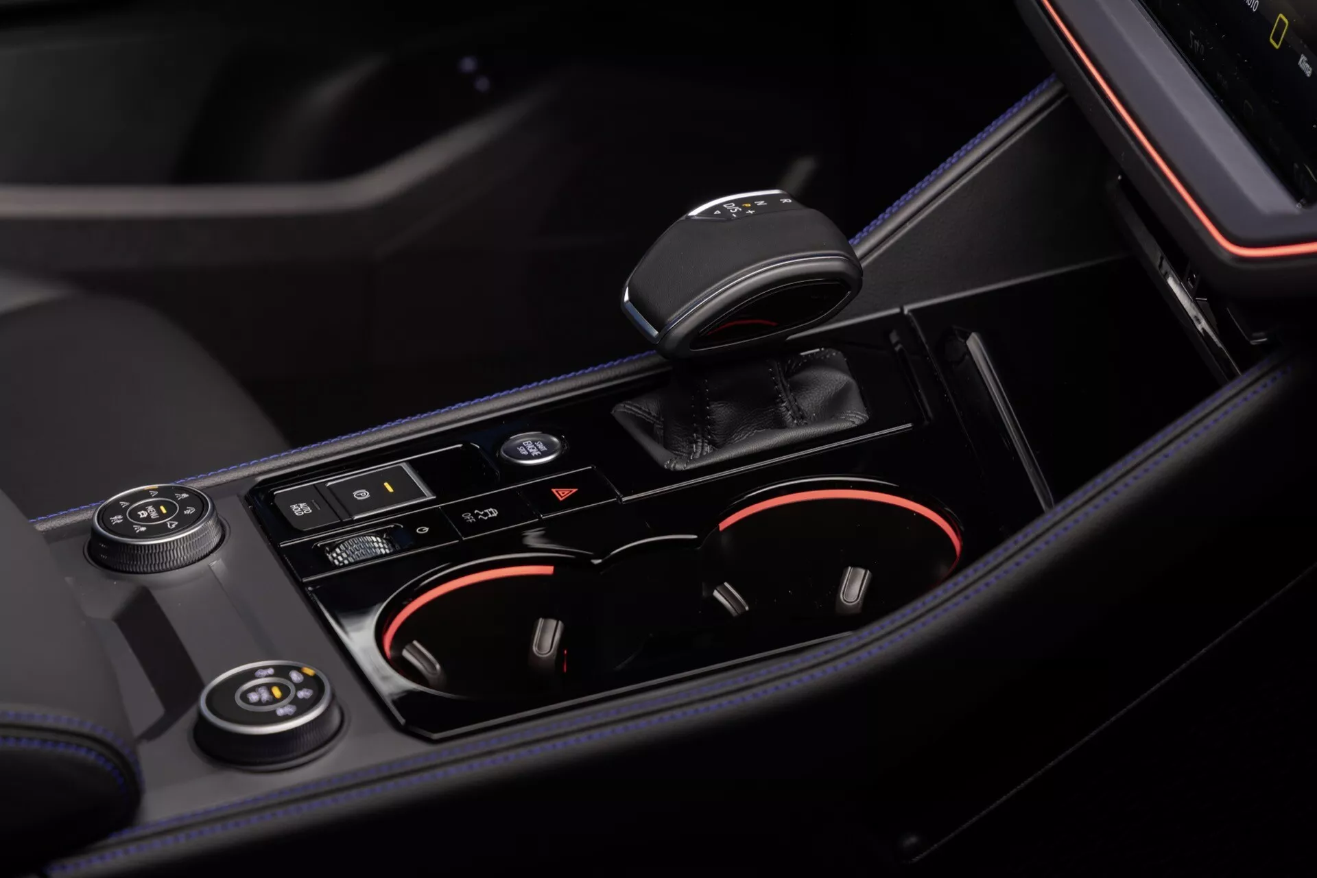 Intérieur du VW Touareg III Facelift 2023 présentant une vue rapprochée de la console centrale avec levier de vitesse automatique, porte-gobelets illuminés, boutons de contrôle pour les fonctionnalités de conduite et boutons de commande de climatisation, mettant en avant des matériaux de haute qualité et un schéma de couleurs noir