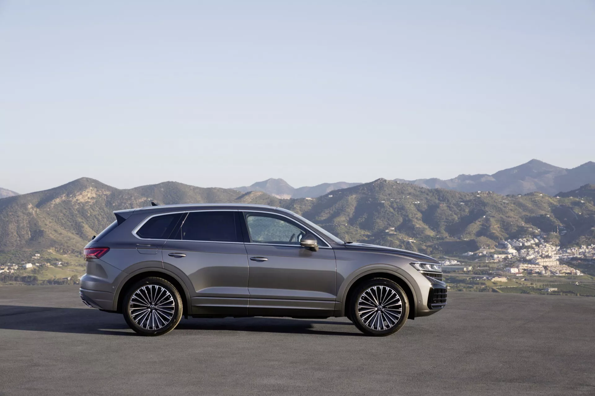VW Touareg III restylé 2023 en profil latéral garé sur une surface plane avec un arrière-plan montagneux sous un ciel clair, présentant son design moderne de SUV, ses jantes en alliage stylées et son toit panoramique.
