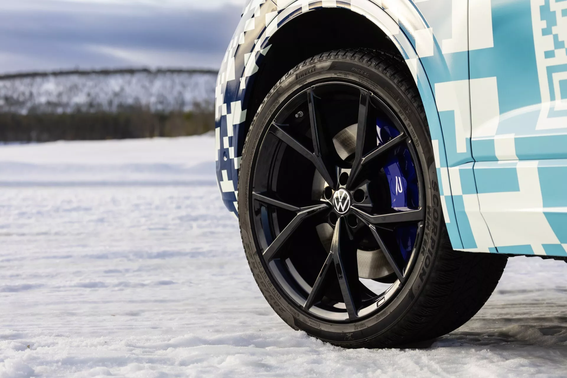 Vue rapprochée de la roue avant du VW Touareg III Facelift 2023 avec jante noire à multiples branches, logo Volkswagen et étriers de frein bleus sur une surface enneigée, mettant en avant les caractéristiques de performance et les tests hivernaux