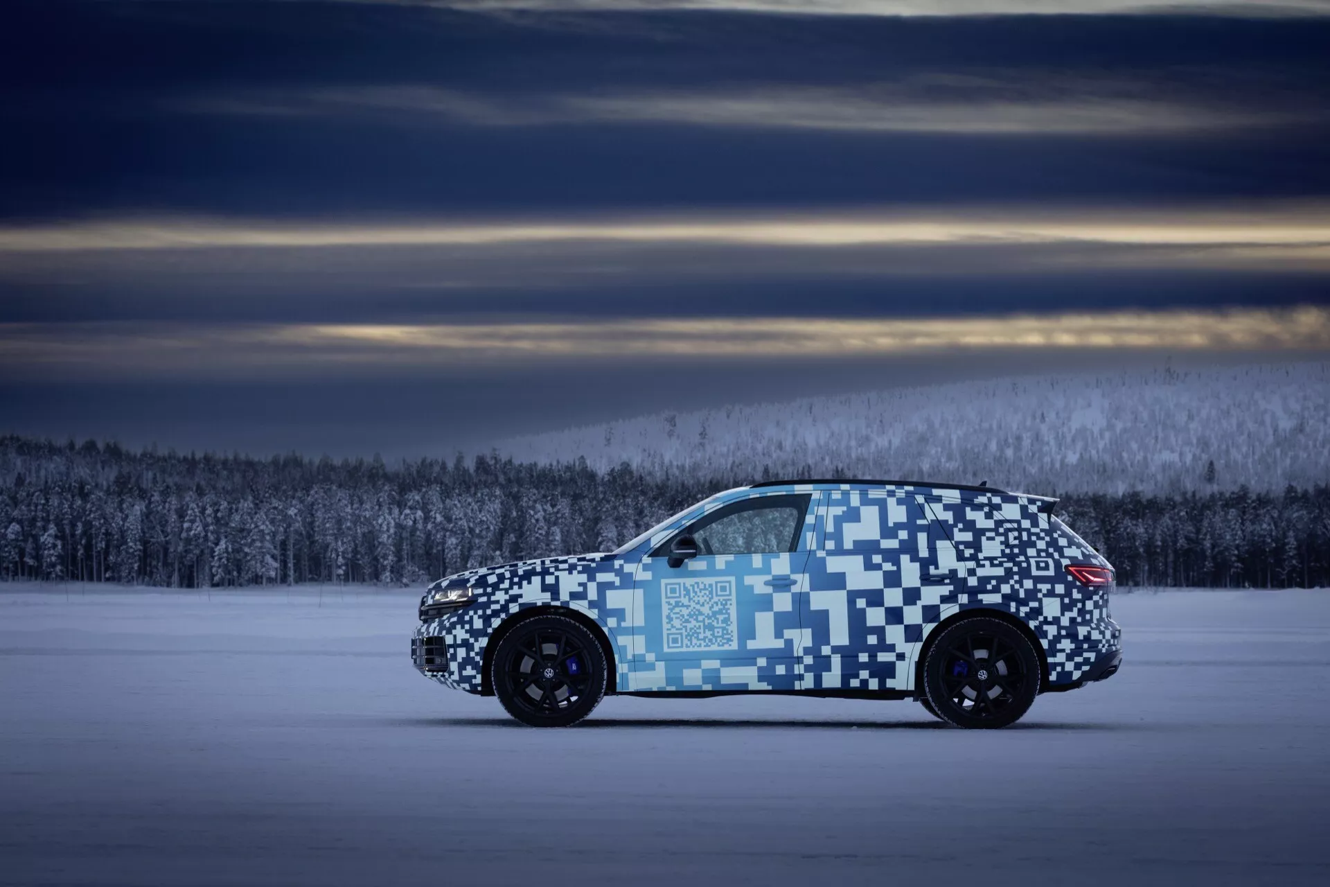 Vue de profil du VW Touareg 2023 (III facelift) présentant un camouflage pixelisé distinctif sur un terrain couvert de neige au crépuscule, mettant en évidence sa phase de test dans un paysage hivernal avec un design de roues à multiples branches visible.