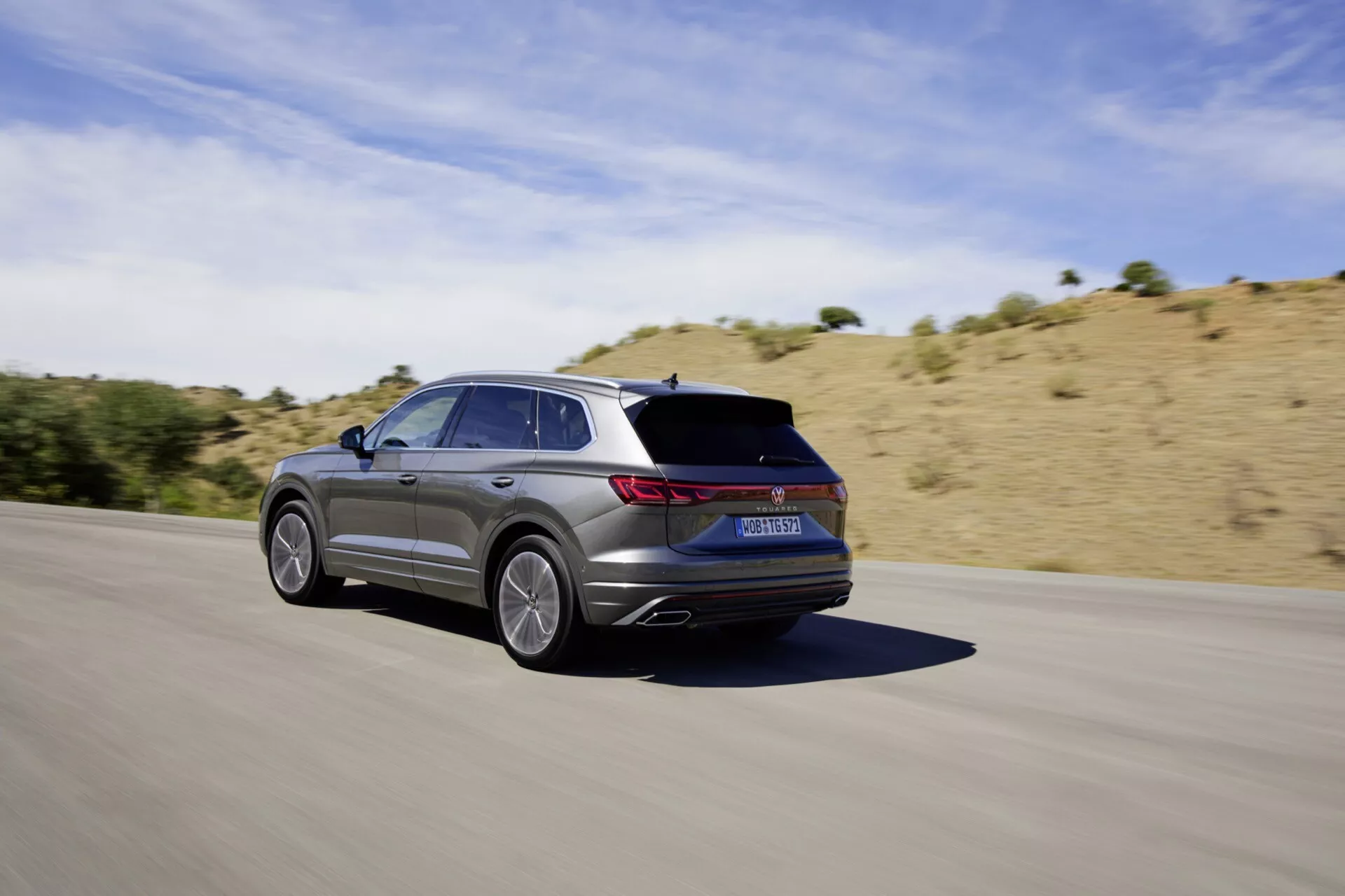 VW Touareg III facelift 2023 en mouvement sur une route en asphalte, capturé depuis un angle arrière trois quarts montrant son design élégant, ses jantes en alliage à cinq branches, sa finition métallique, ses feux arrière LED, ses sorties d'échappement intégrées et son esthétique sportive contre un paysage sec et vallonné sous un ciel ensoleillé clair