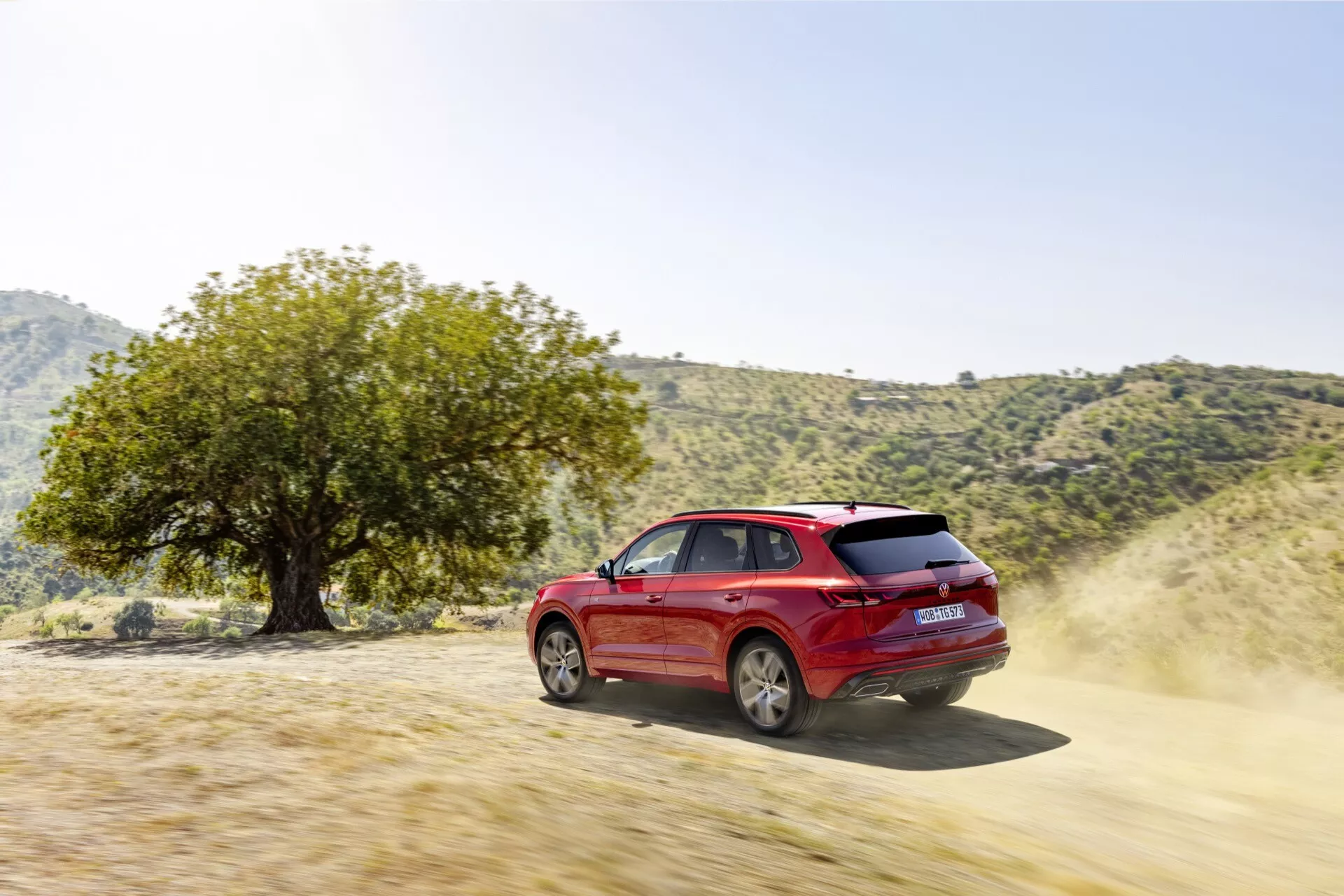 SUV rouge Volkswagen Touareg III Facelift 2023 mettant en évidence les capacités tout-terrain sur un sentier de terre, vu sous un angle arrière de trois quarts soulignant son design dynamique, jantes, portes latérales, fenêtres latérales et arrière, avec de la poussière indiquant un mouvement et des caractéristiques de performance comme la puissance en chevaux et la capacité 4x4, dans un cadre naturel extérieur suggérant l'aventure et la polyvalence tout-terrain.