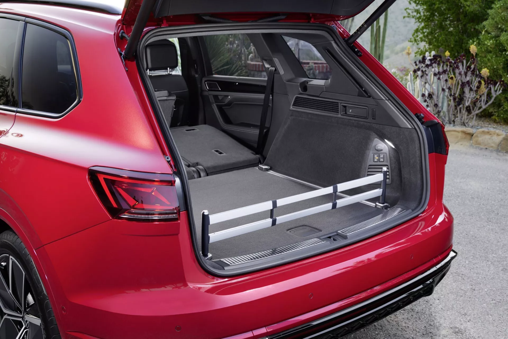 Image détaillée de la Volkswagen Touareg III restylée 2023 montrant l'espace de chargement spacieux avec un plancher plat de chargement, un revêtement protecteur et des compartiments de rangement intégrés depuis une vue arrière droite en angle de trois quarts, disposant d'une prise 12V, de barres de chargement ajustables et de sièges arrière partiellement rabattus pour agrandir l'espace, soulignée par l'extérieur rouge du véhicule et l'aileron arrière sportif.